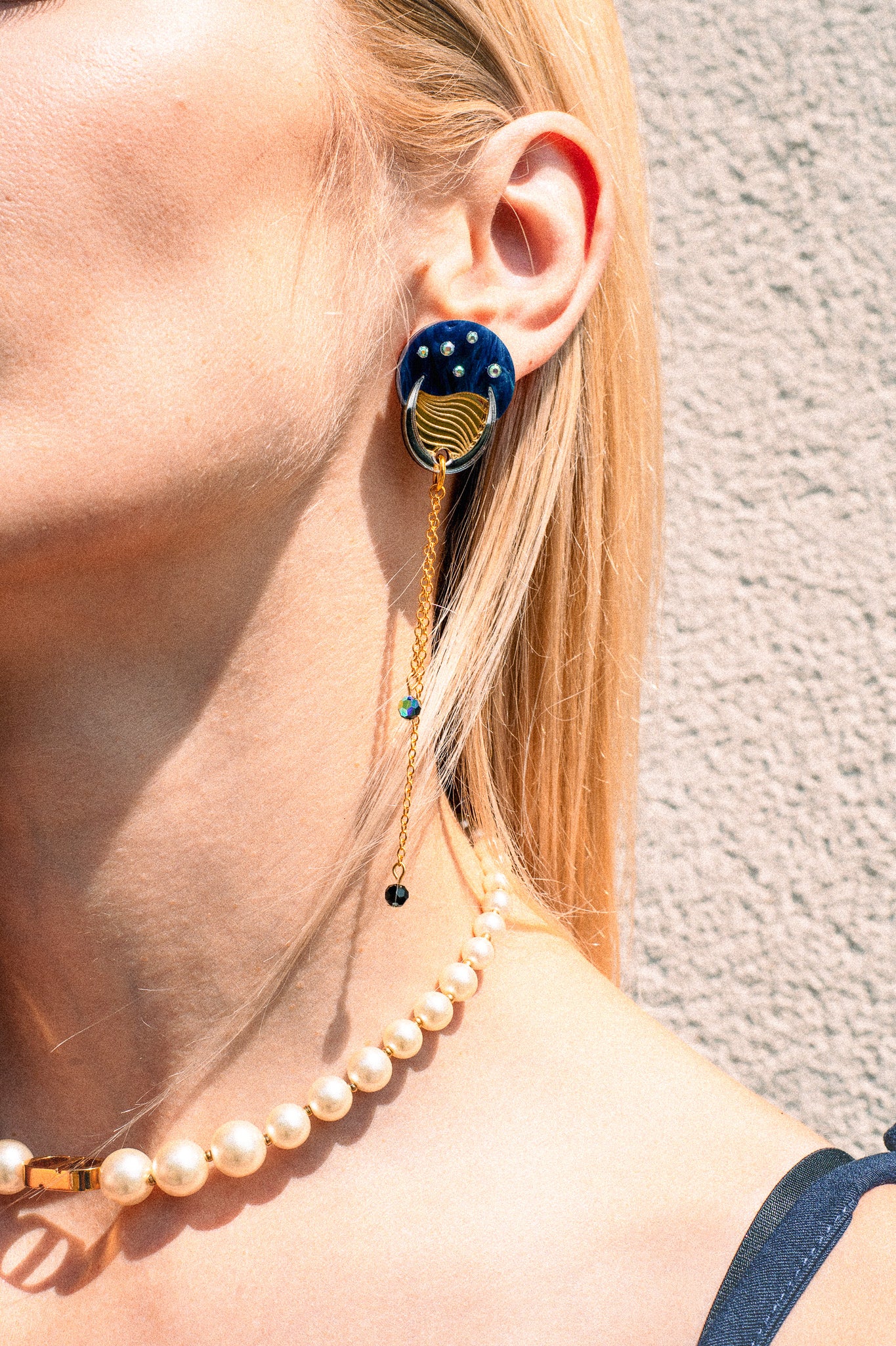 A close-up of a person wearing a blue and gold statement earring with dangling chains that evoke the shimmer of the night sky, paired with a pearl necklace. The person has blonde hair and light skin, and stands against a textured beige wall.