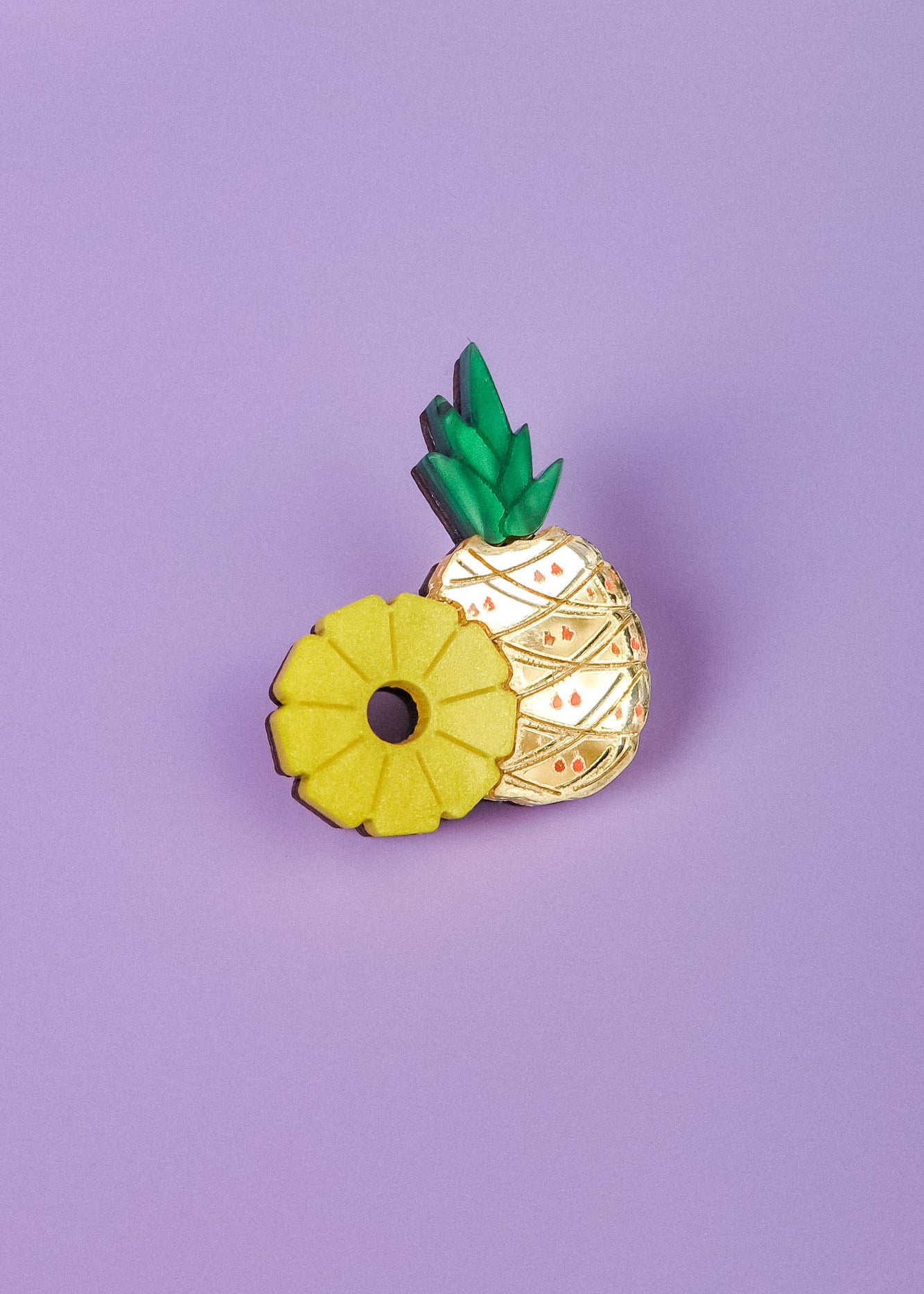 A handcrafted Pineapple Pin with green leaves and gold detailing stands on a light purple background, next to a yellow ring-shaped accessory resembling a sliced pineapple.
