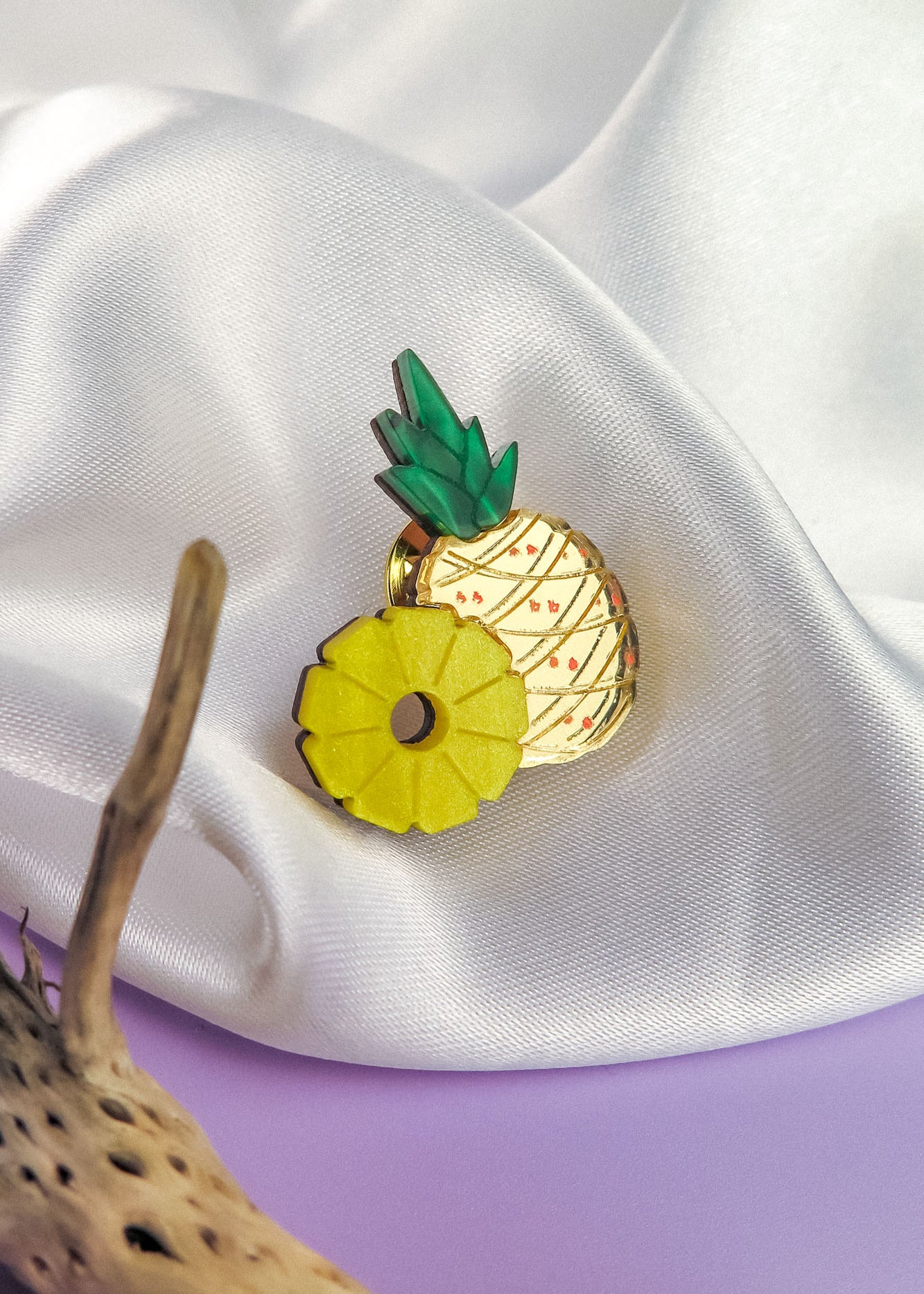 A handcrafted Pineapple Pin accessory with green leaves and a yellow ring rests on white satin fabric, beside textured brown driftwood on a purple surface.