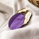 A purple and gold acrylic brooch shaped like a plum with two leaves rests on a soft, white fabric near a woven basket.