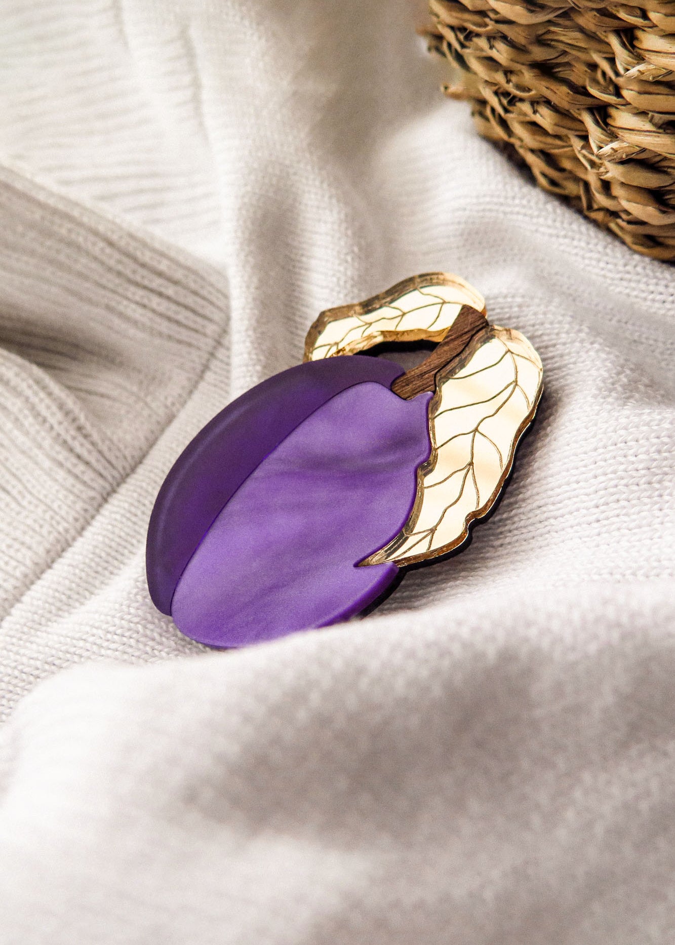 A purple and gold acrylic brooch shaped like a plum with two leaves rests on a soft, white fabric near a woven basket.