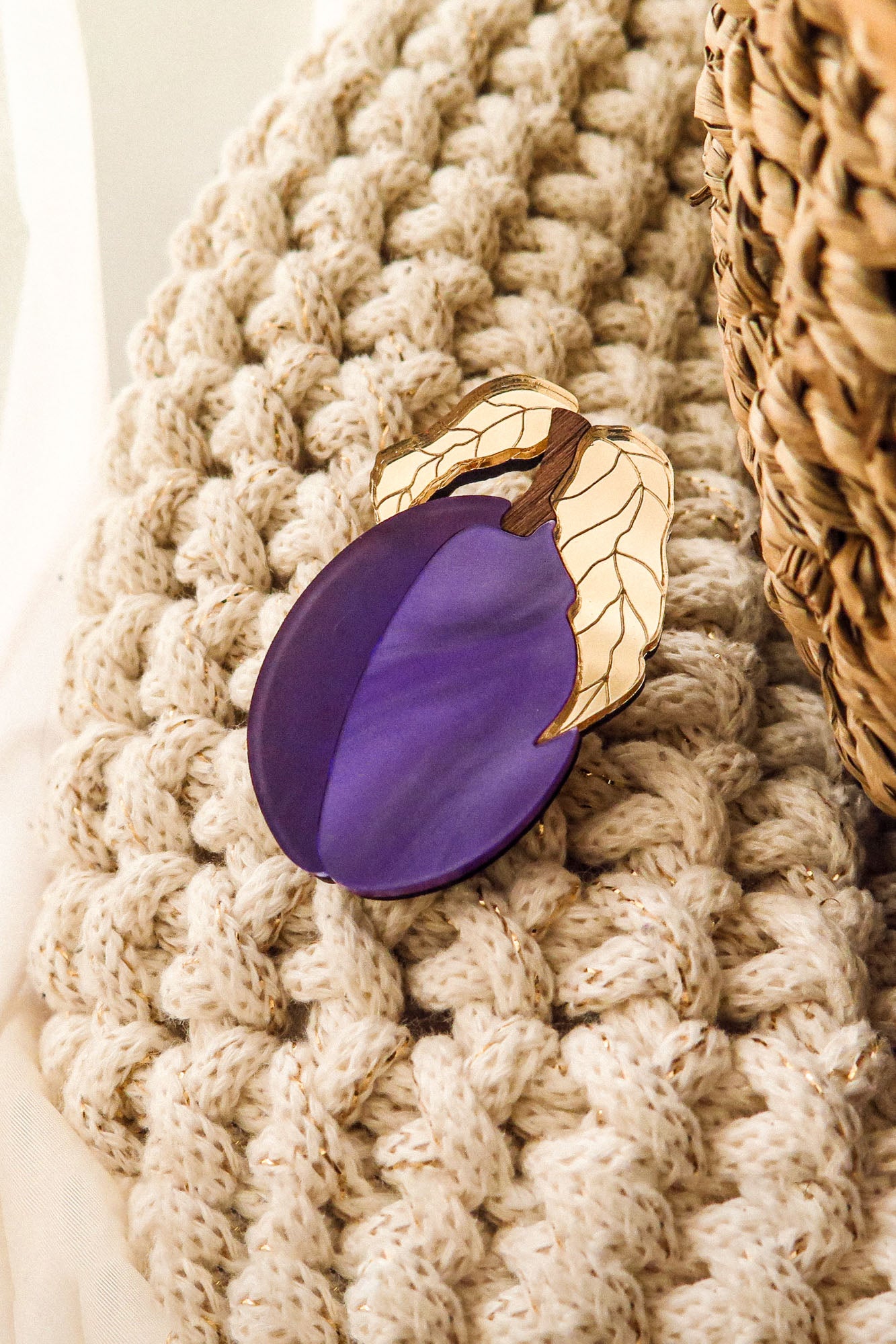 A purple enamel eggplant brooch with gold-outlined leaves rests on a chunky, cream-colored knit blanket beside a woven basket—crafted by a family business.