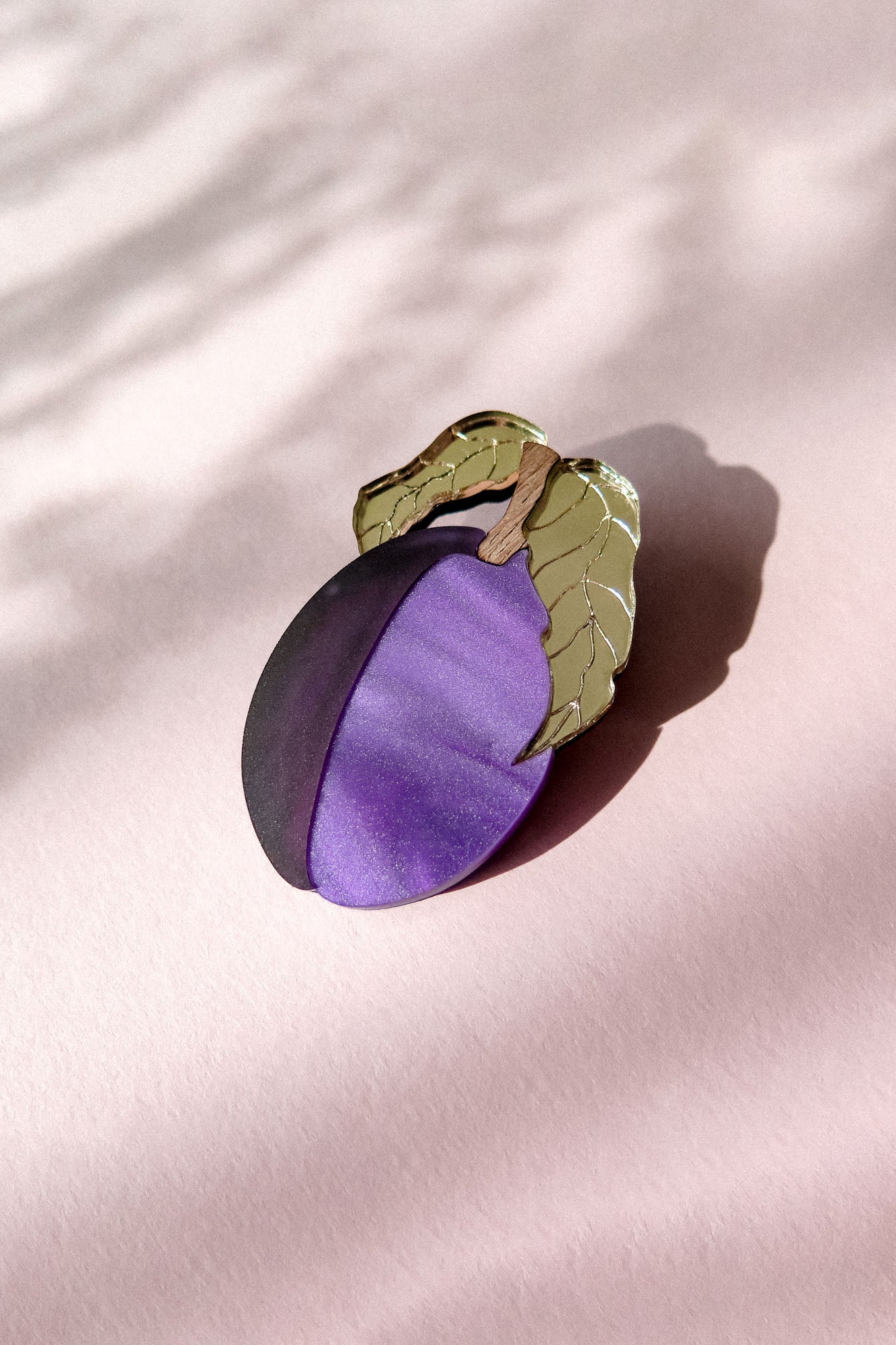 A decorative brooch shaped like a plum with two green leaves, crafted from acrylic and recycled wood, rests on a pale pink surface with soft shadows.