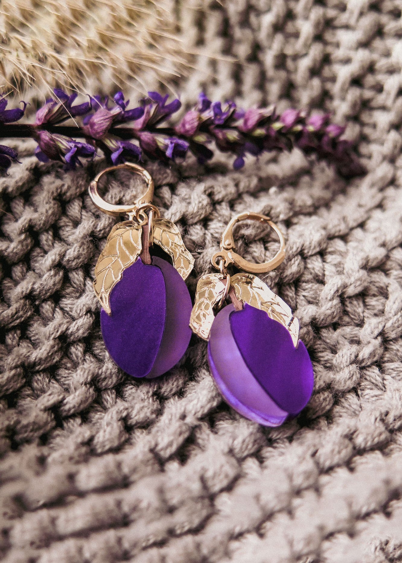 Pair of gold and purple plum earrings with textured gold accents, displayed on a knitted beige fabric with a sprig of purple flowers alongside. These handcrafted earrings add a touch of elegance to any outfit.