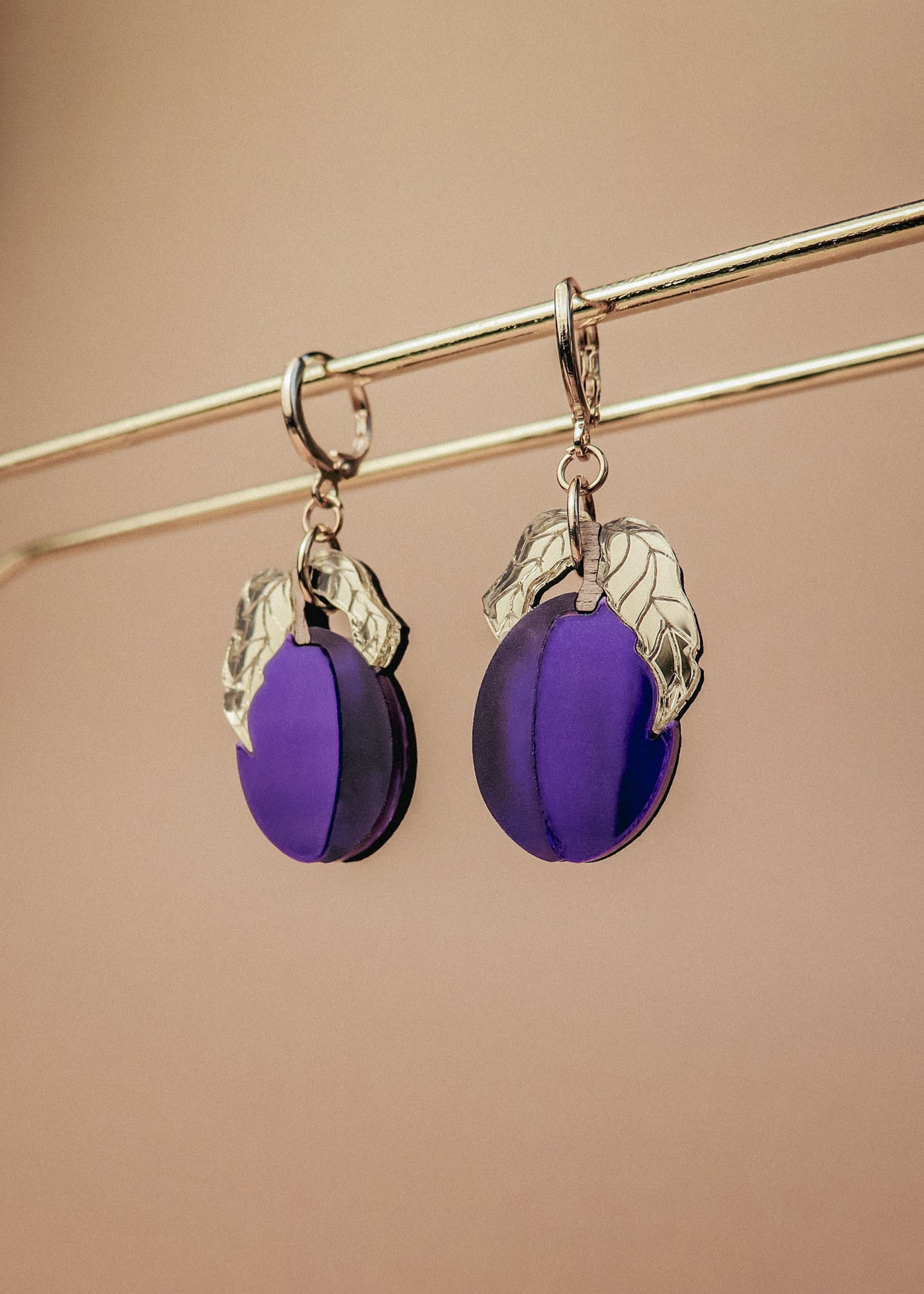 A pair of elegant purple plum earrings with silver leaf accents hang from a gold-colored rod against a beige background.