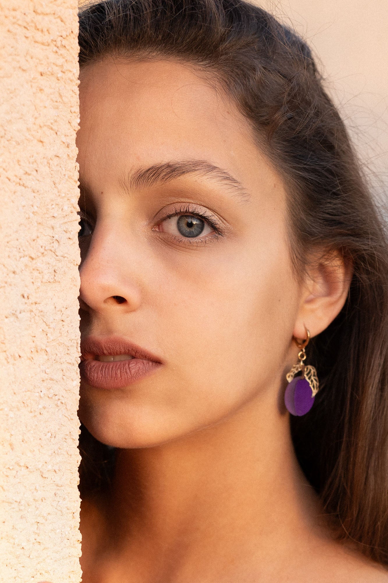 A woman with long brown hair and elegant purple plum earrings looks directly at the camera, standing next to a textured beige wall, with soft natural light highlighting her face.