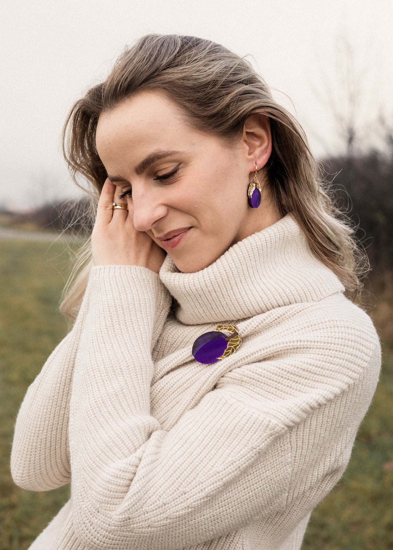 A woman wearing a cream-colored turtleneck sweater stands outdoors, smiling gently with eyes closed. She has wavy hair and wears matching purple jewelry—a handcrafted brooch and earrings. The grassy background is slightly blurred.