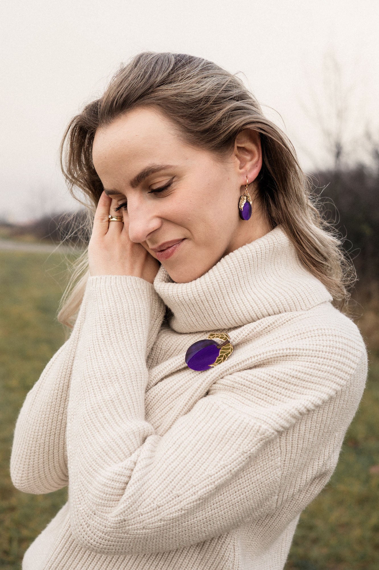 A woman wearing a cream-colored turtleneck sweater stands outdoors, smiling gently with eyes closed. She has wavy hair and wears matching purple jewelry—a handcrafted brooch and earrings. The grassy background is slightly blurred.