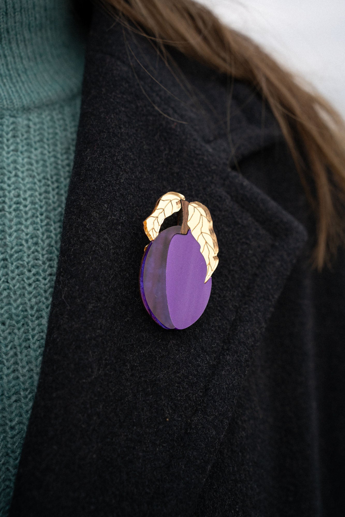 A person wearing a dark coat with an elegant brooch shaped like a purple fruit and two gold-colored leaves pinned to the lapel. The person is also wearing a teal knit sweater.