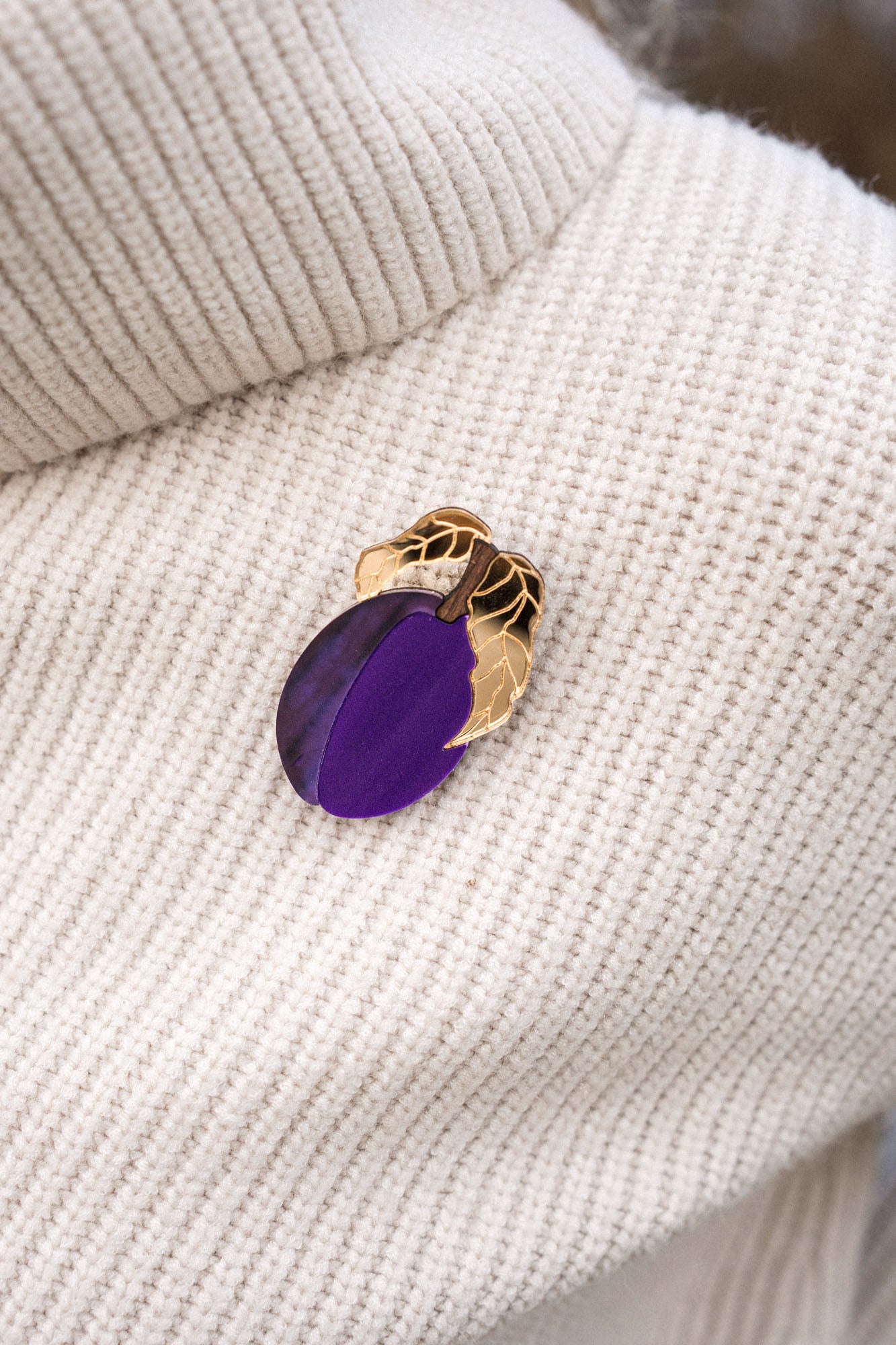 A handcrafted brooch shaped like a gold and purple eggplant with two gold leaves is pinned to a cream-colored ribbed sweater.