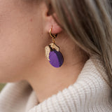 Close-up of a person wearing elegant purple plum earrings shaped like fruit with gold leaves. The person has light brown hair and is wearing a cream-colored, ribbed turtleneck sweater.