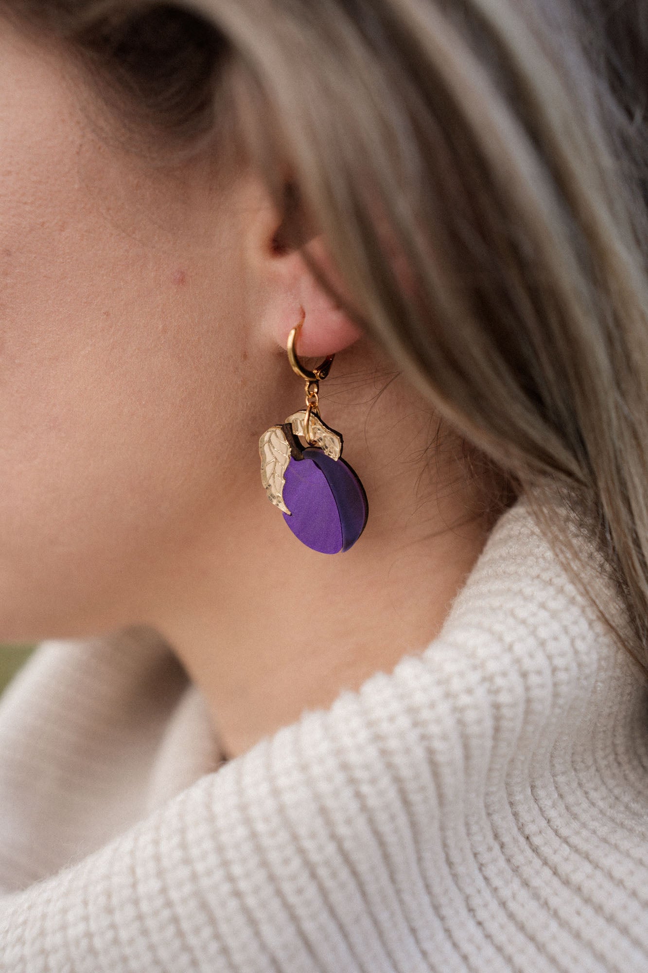 Close-up of a person wearing elegant purple plum earrings shaped like fruit with gold leaves. The person has light brown hair and is wearing a cream-colored, ribbed turtleneck sweater.