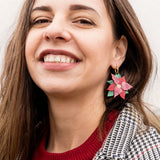 A woman with long brown hair smiles, wearing a red sweater and checkered coat. She showcases festive jewelry with large Poinsettia Statement Hoops, holiday earrings shaped like red and green poinsettias. The background is softly blurred.