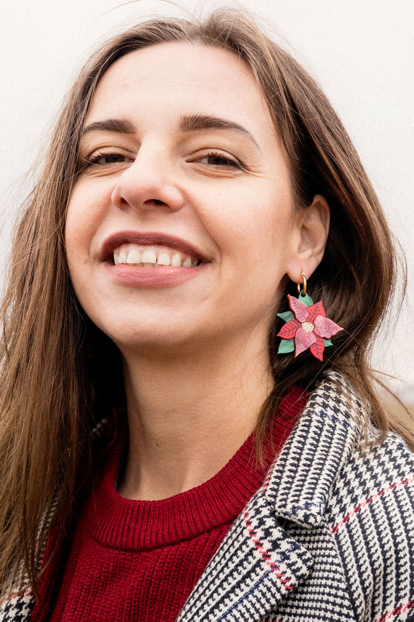 A woman with long brown hair smiles, wearing a red sweater and checkered coat. She showcases festive jewelry with large Poinsettia Statement Hoops, holiday earrings shaped like red and green poinsettias. The background is softly blurred.