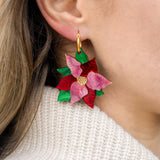 Close-up of a person's ear wearing festive jewelry: colorful, flower-shaped Poinsettia Statement Hoops with red and pink petals, green leaves, and a gold center, paired with a cream knit sweater.