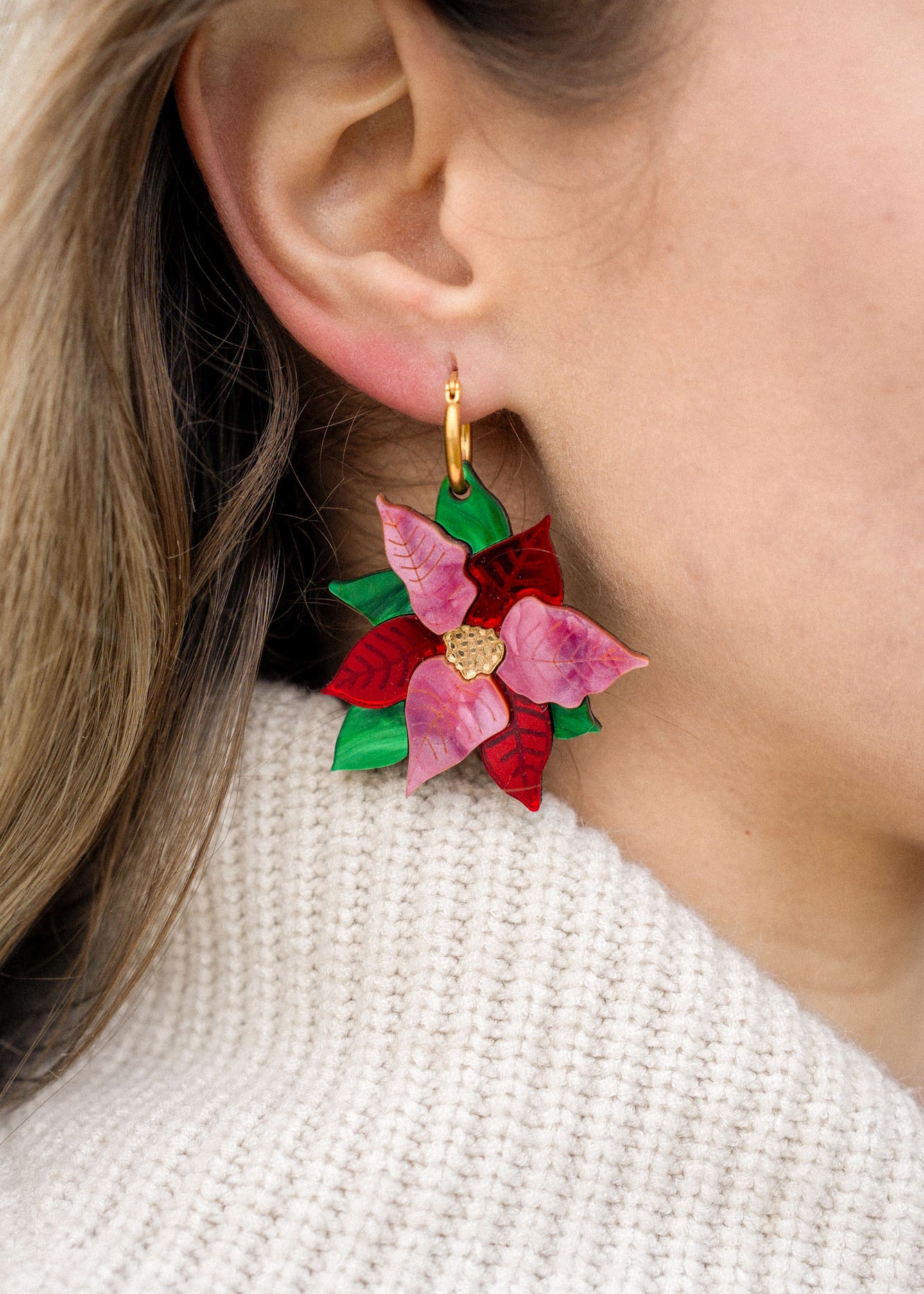 Close-up of a person's ear wearing festive jewelry: colorful, flower-shaped Poinsettia Statement Hoops with red and pink petals, green leaves, and a gold center, paired with a cream knit sweater.