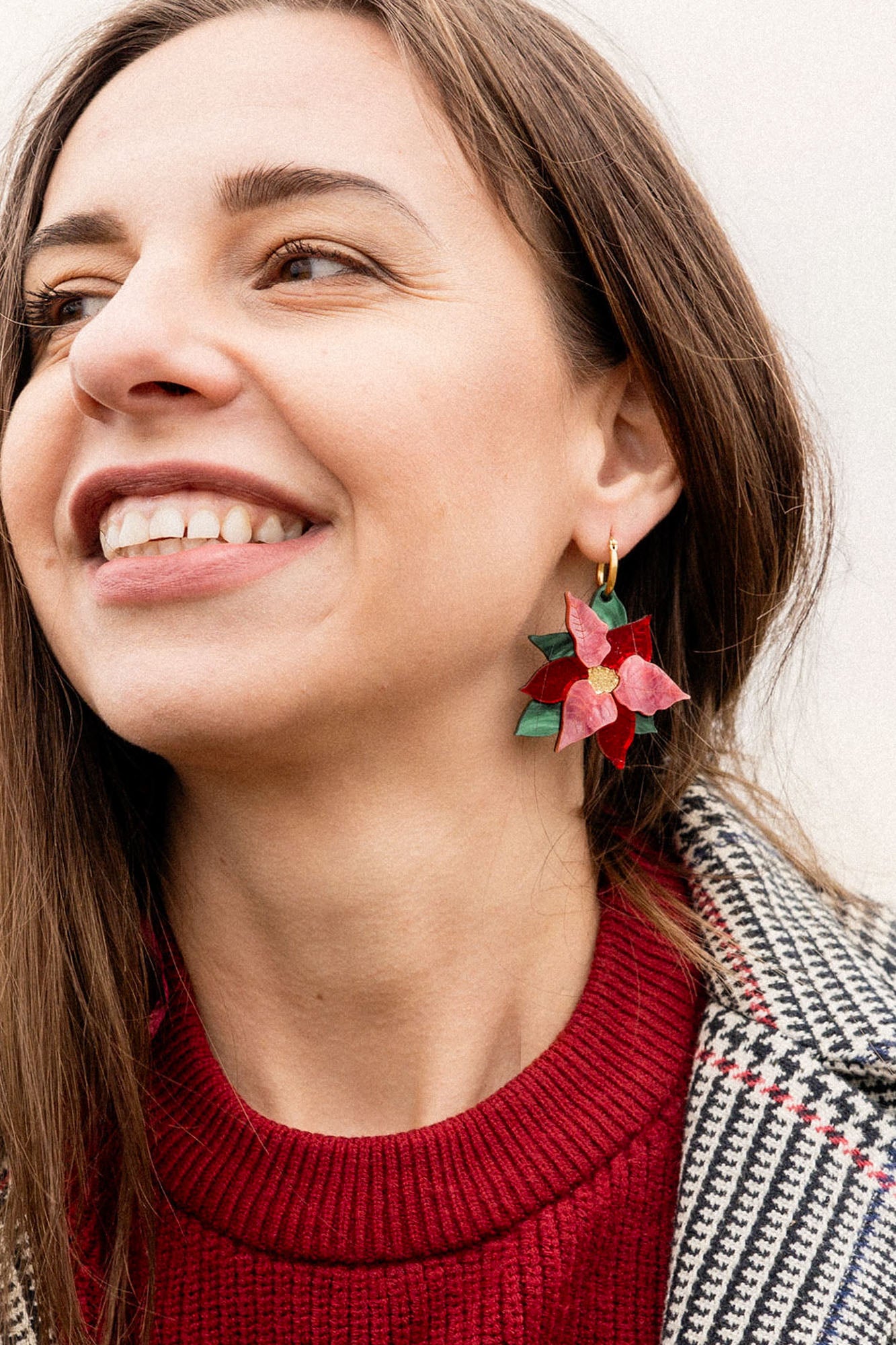 A woman smiling, wearing a red sweater and a plaid coat, shows off her festive jewelry—a poinsettia-shaped earring with red petals and green leaves, perfect as holiday earrings.
