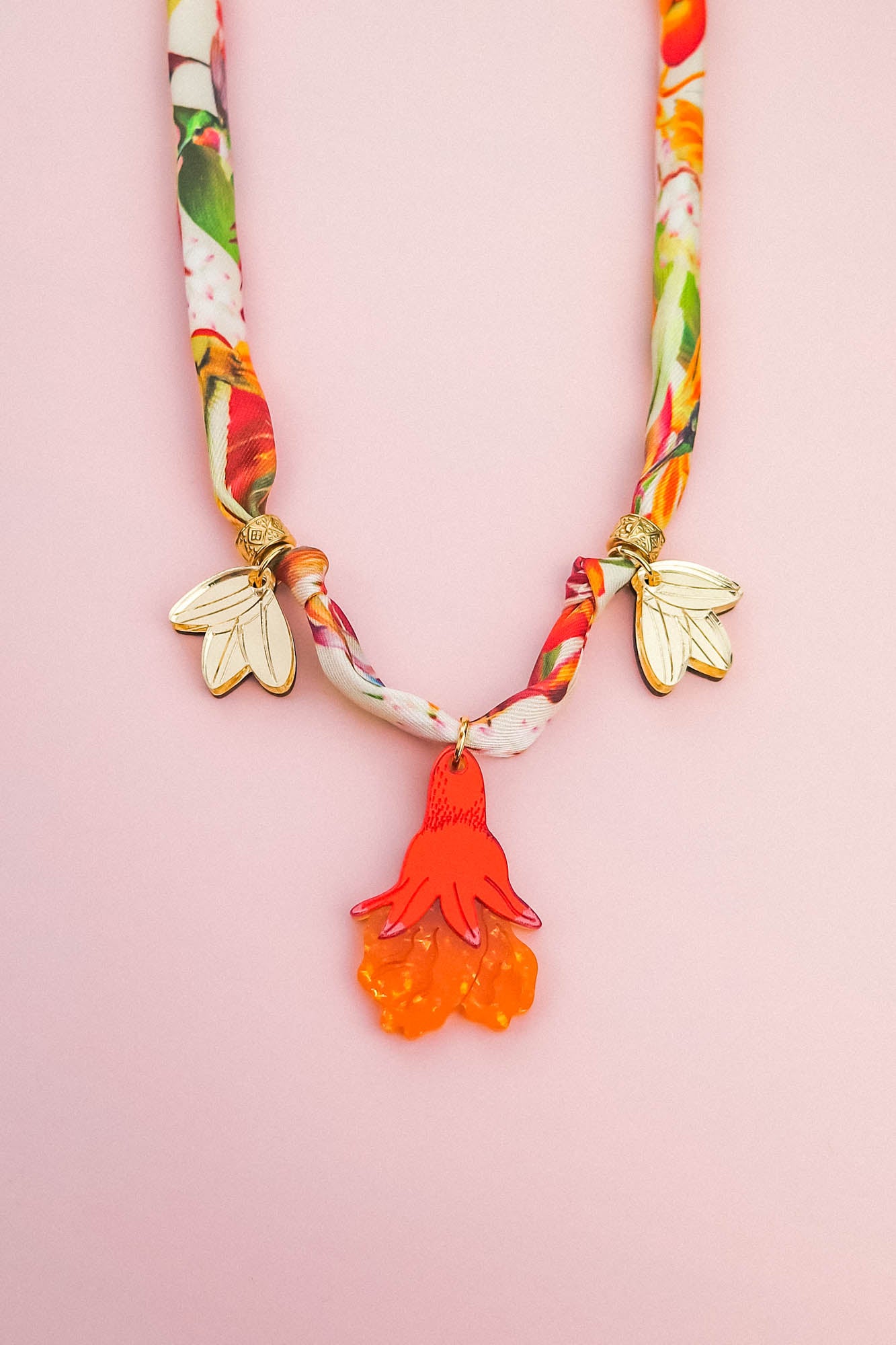 A colorful handmade necklace with a floral-patterned fabric band, two gold flower charms, and a central pendant shaped like an orange and red flower—perfect as a unique pomegranate flower necklace—displayed on a light pink background.