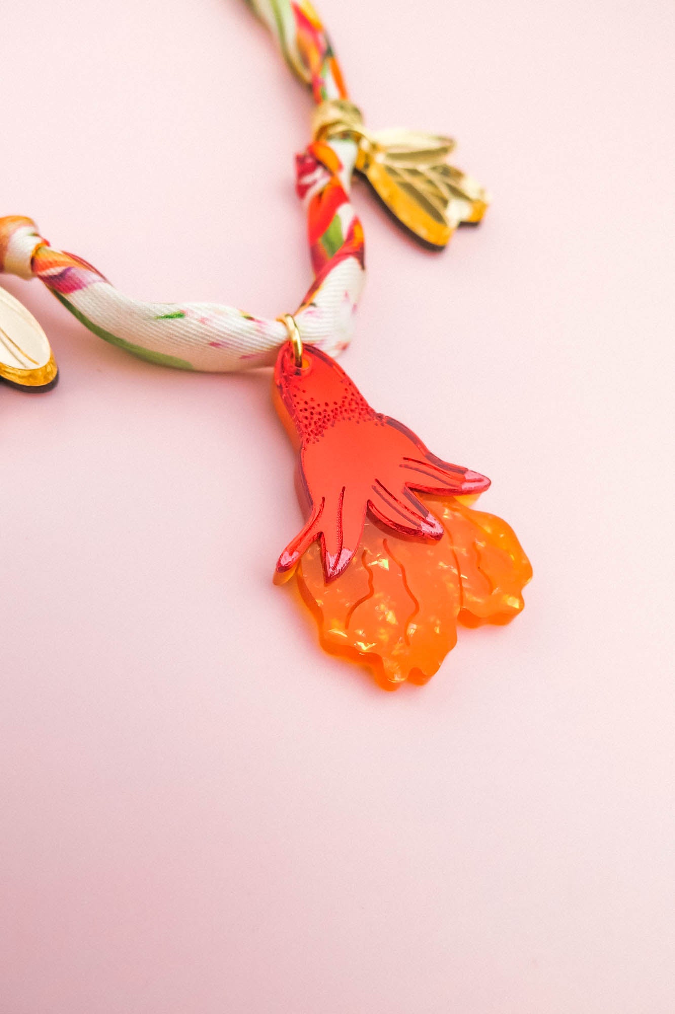 A colorful handmade necklace with a twisted fabric band and gold leaves features an orange, hand-shaped pendant with textured details, set against a light pink background.