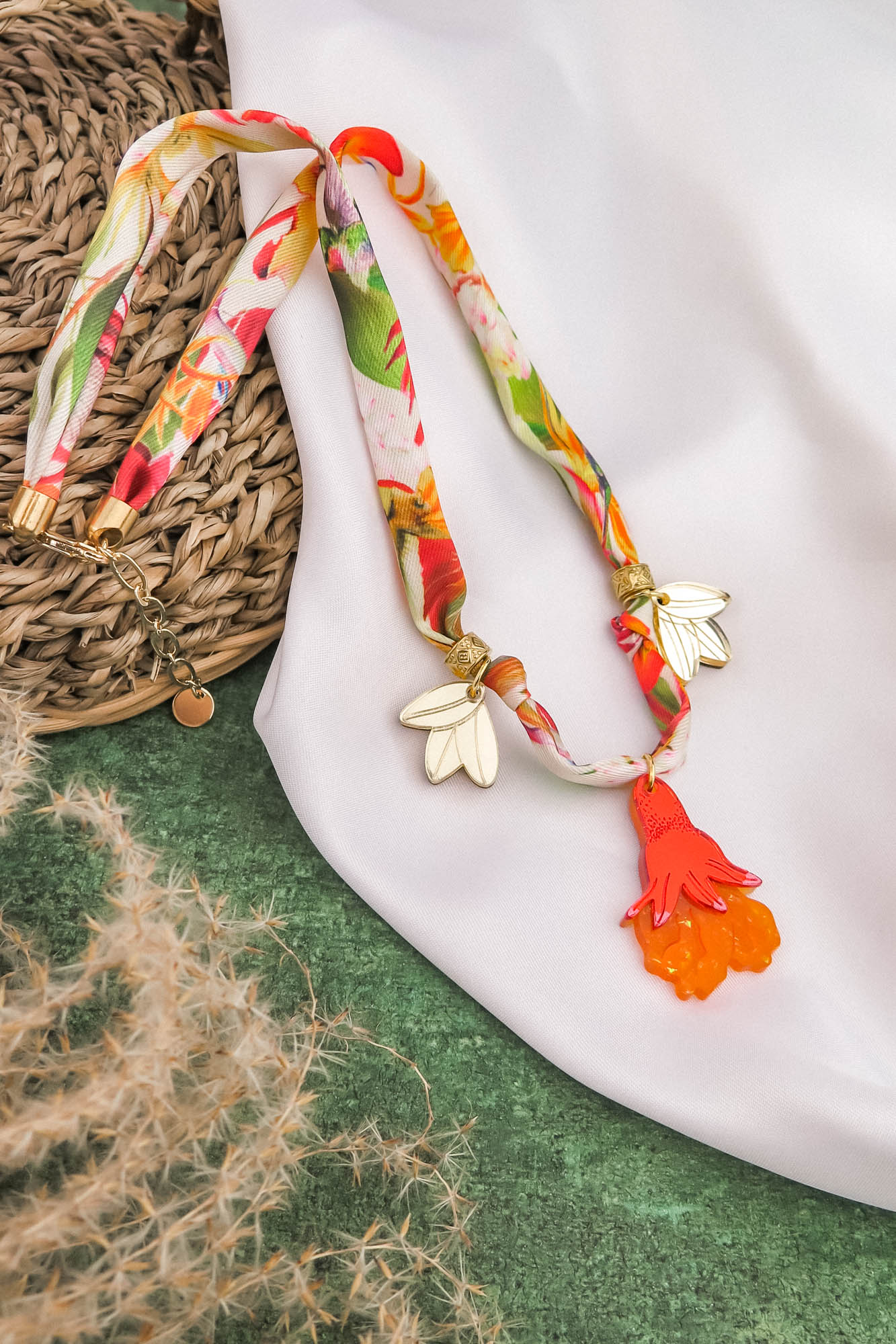 A colorful handmade necklace with floral patterns, featuring two gold and white wing-shaped charms and a bright orange hand-shaped pendant, is displayed on a white cloth and green textured background.