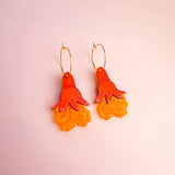 A pair of Little Turtle Hoop Earrings with gold hoops and dangling charms shaped like stylized orange and yellow flowers, displayed on a light pink background. These handcrafted earrings add a cheerful touch to any look.