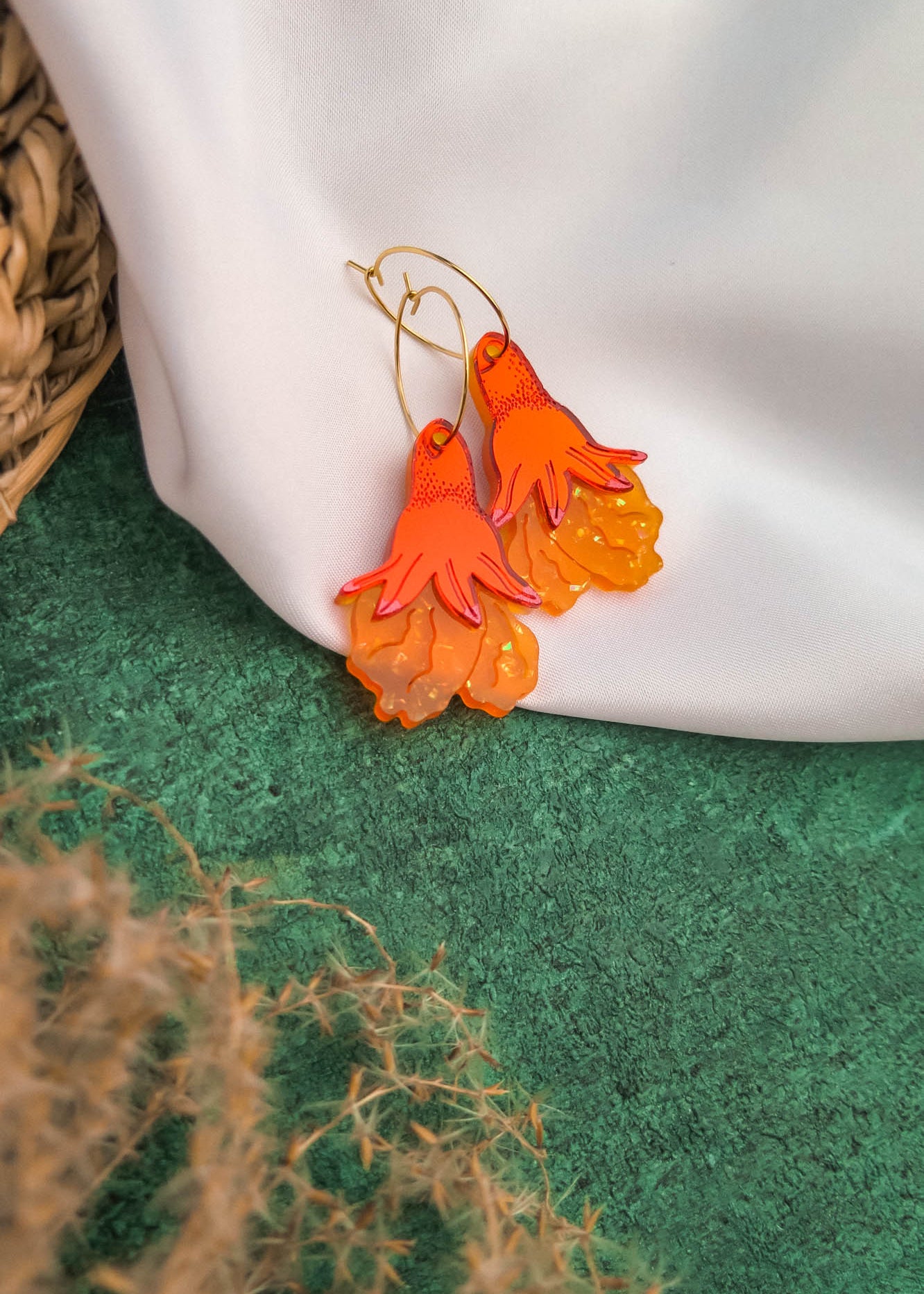 A pair of orange and yellow handcrafted earrings, shaped like stylized flowers or flames, rest on a white fabric backdrop. Green textured ground and part of a woven basket are visible, highlighting these unique gold hoops.