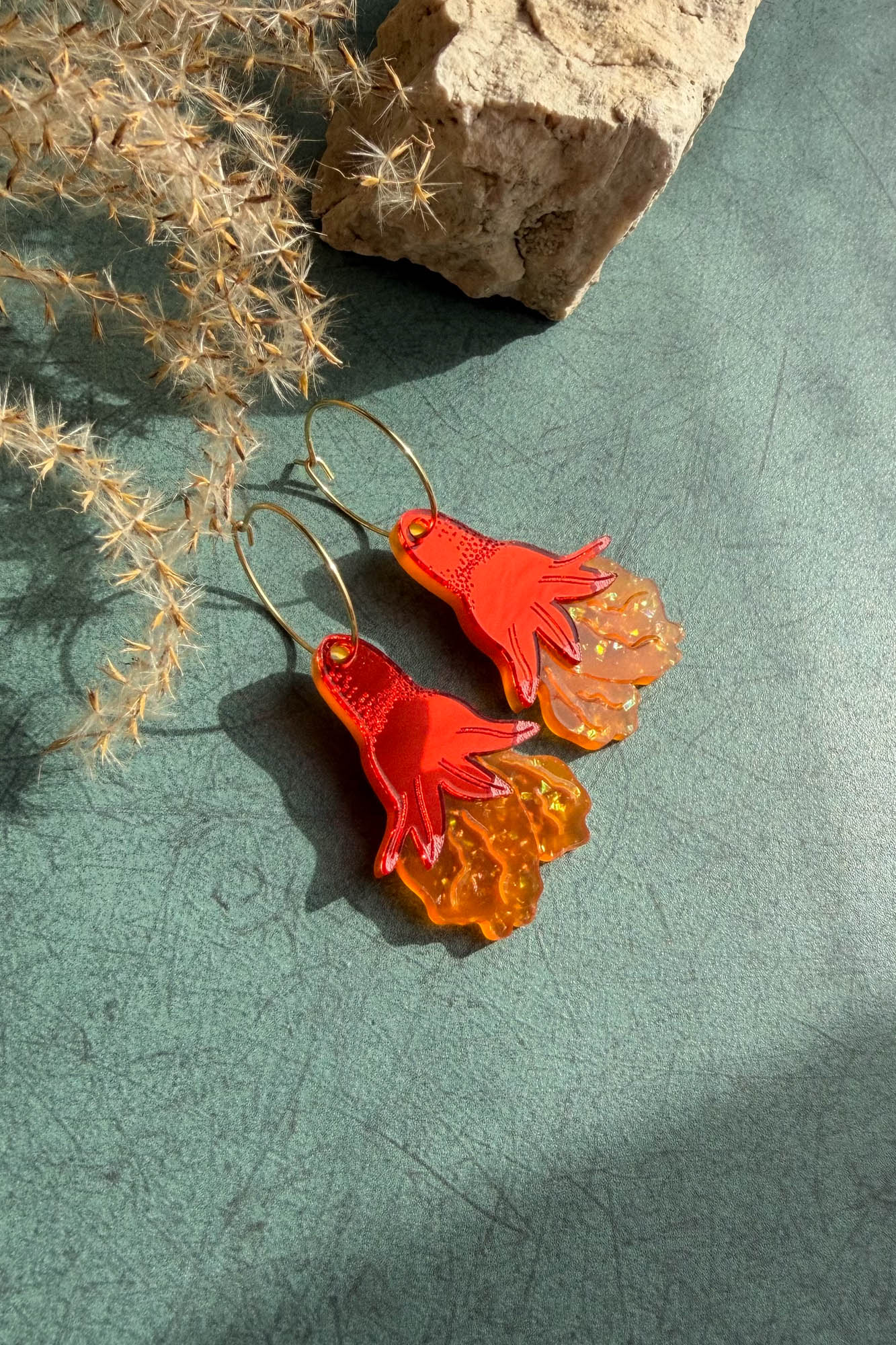 A pair of handcrafted earrings shaped like bright red-orange octopus tentacles with wavy, translucent lower sections, lying on a green surface near dried grass and a beige rock.