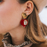 A close-up of a woman's ear wearing a gold stud and a large, handcrafted pomegranate-shaped earring with black leaves. She has a beige beret and a brown coat with a plaid collar. Snowflakes rest on her hair and coat, highlighting bold designs.