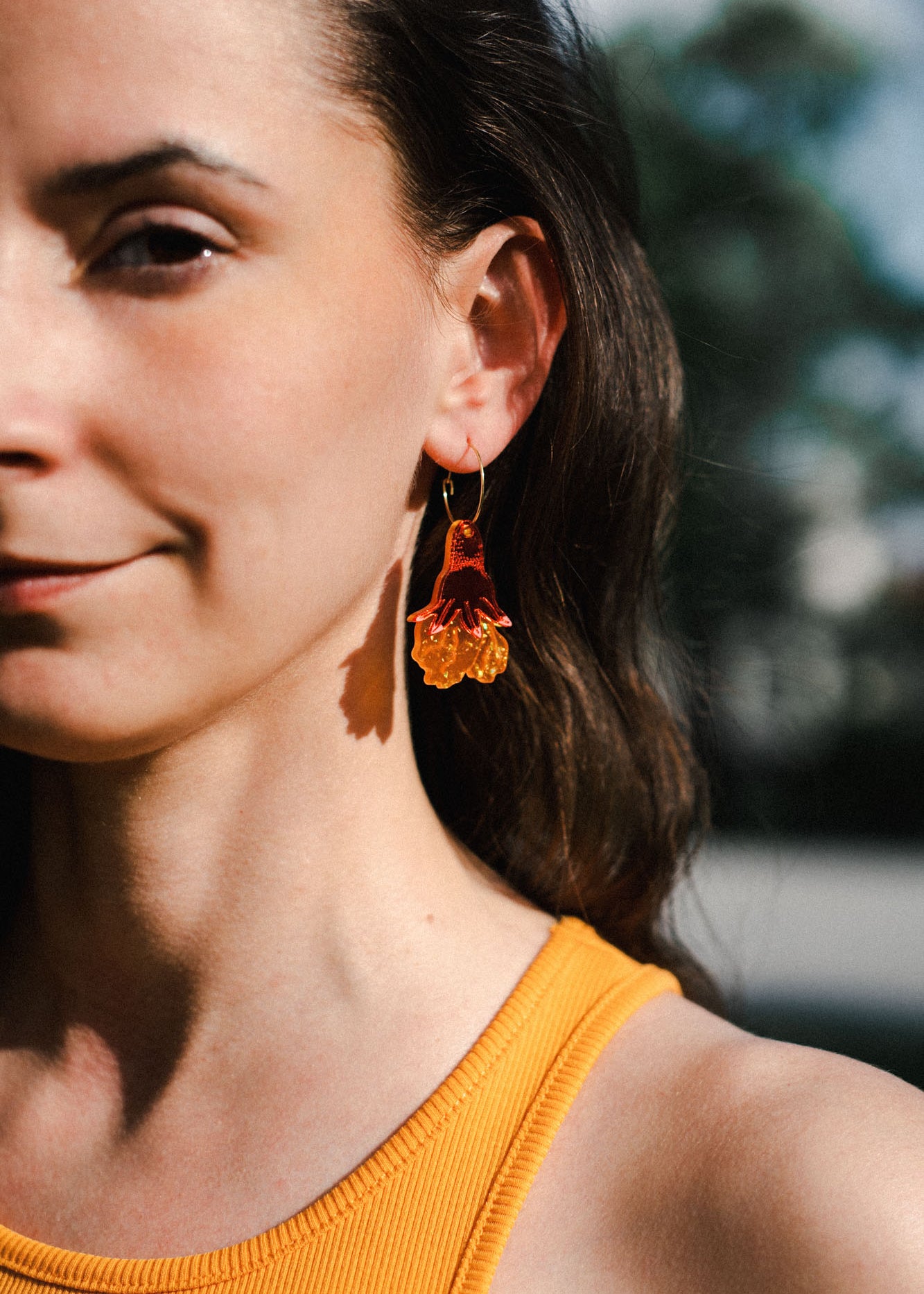 A woman with long brown hair wears handmade floral earrings and a sleeveless yellow top, standing outdoors in natural light. Only the left side of her face and shoulder are visible.