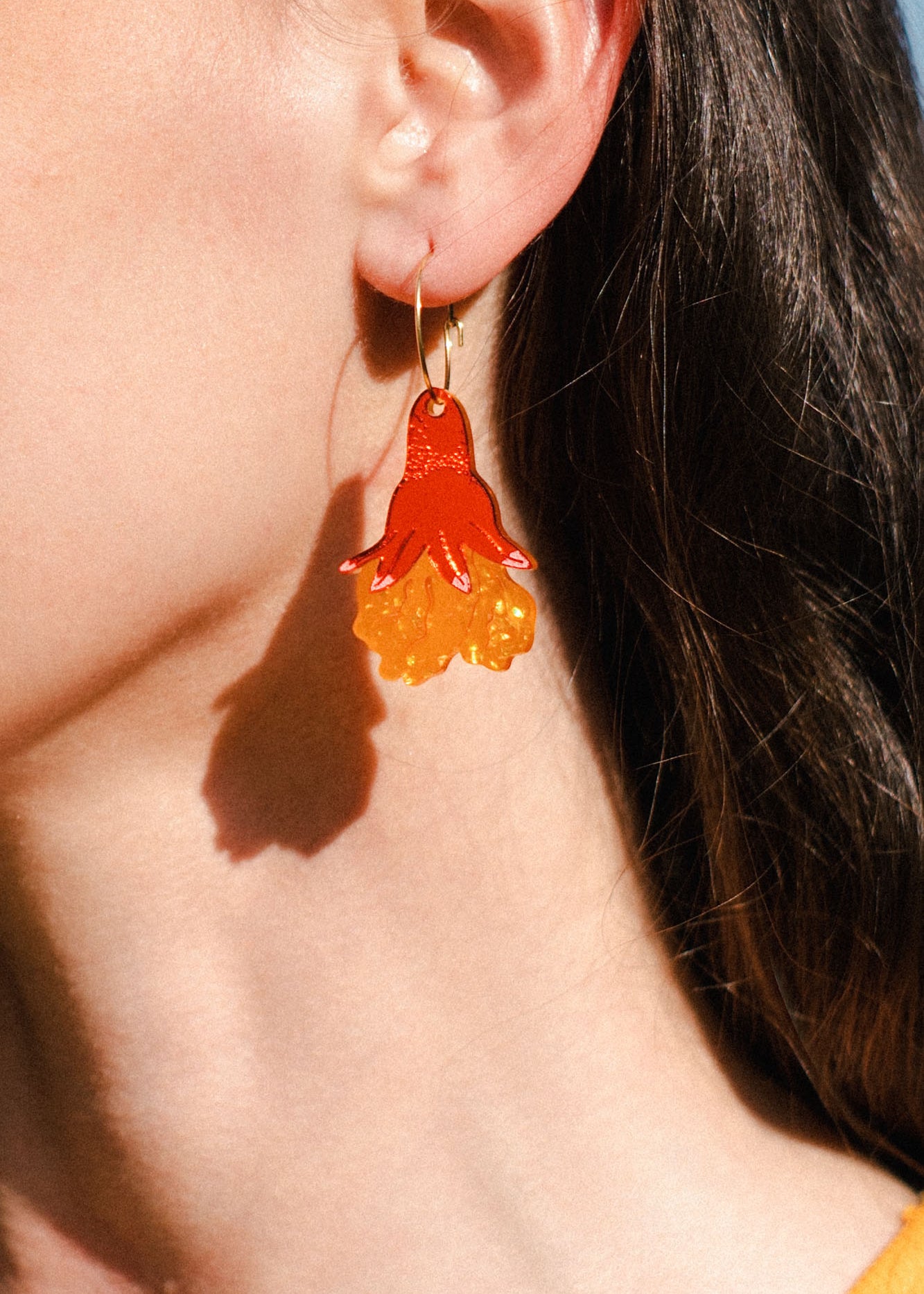 Close-up of a person’s ear wearing bold, handmade floral earrings shaped like a red and yellow jellyfish. The person has long brown hair and stands in bright sunlight, adding summer vibes to these unique accessories.