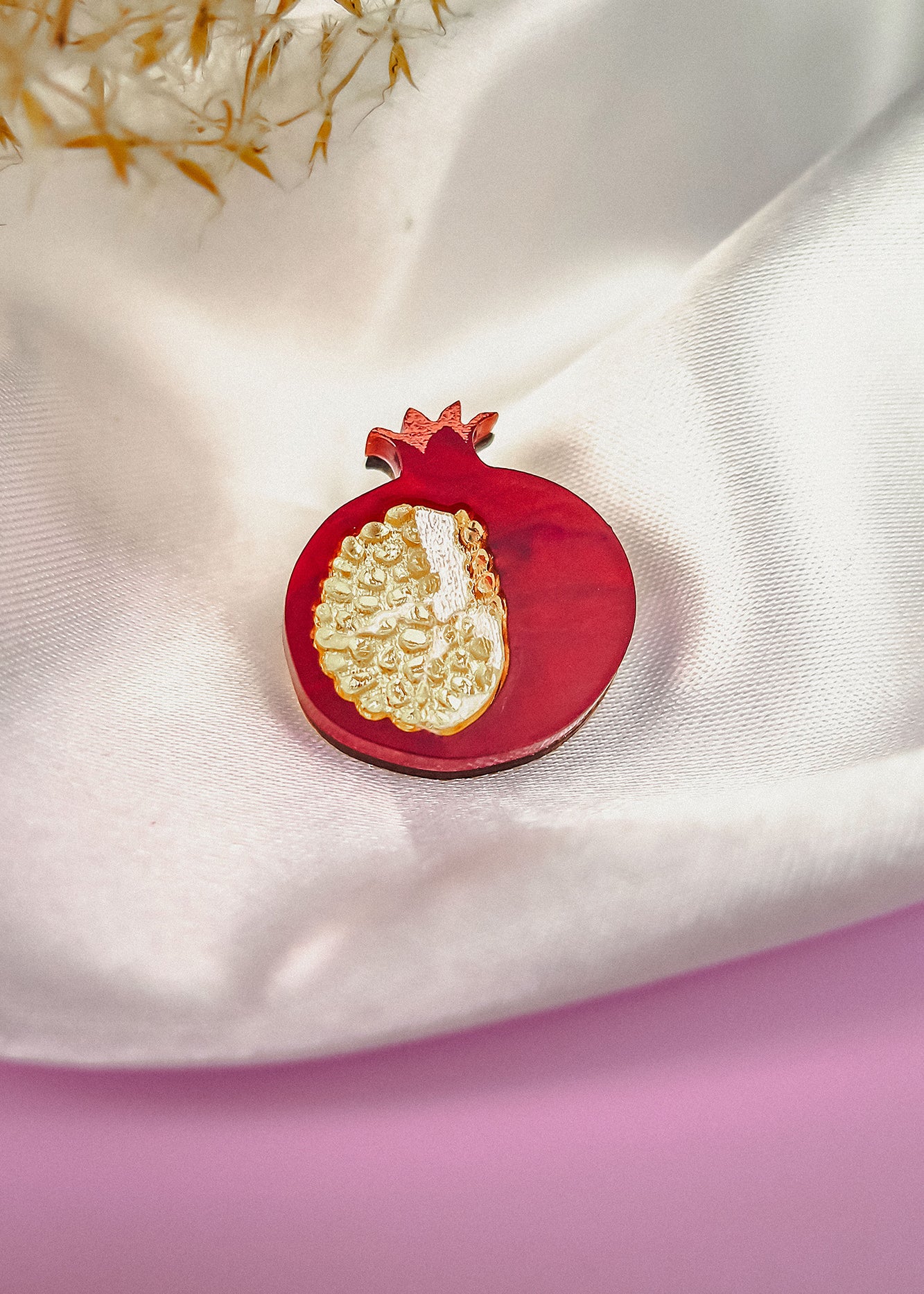 A handcrafted Pomegranate Pin brooch with red and gold details rests on a white satin fabric background, making it an elegant fashion accessory.