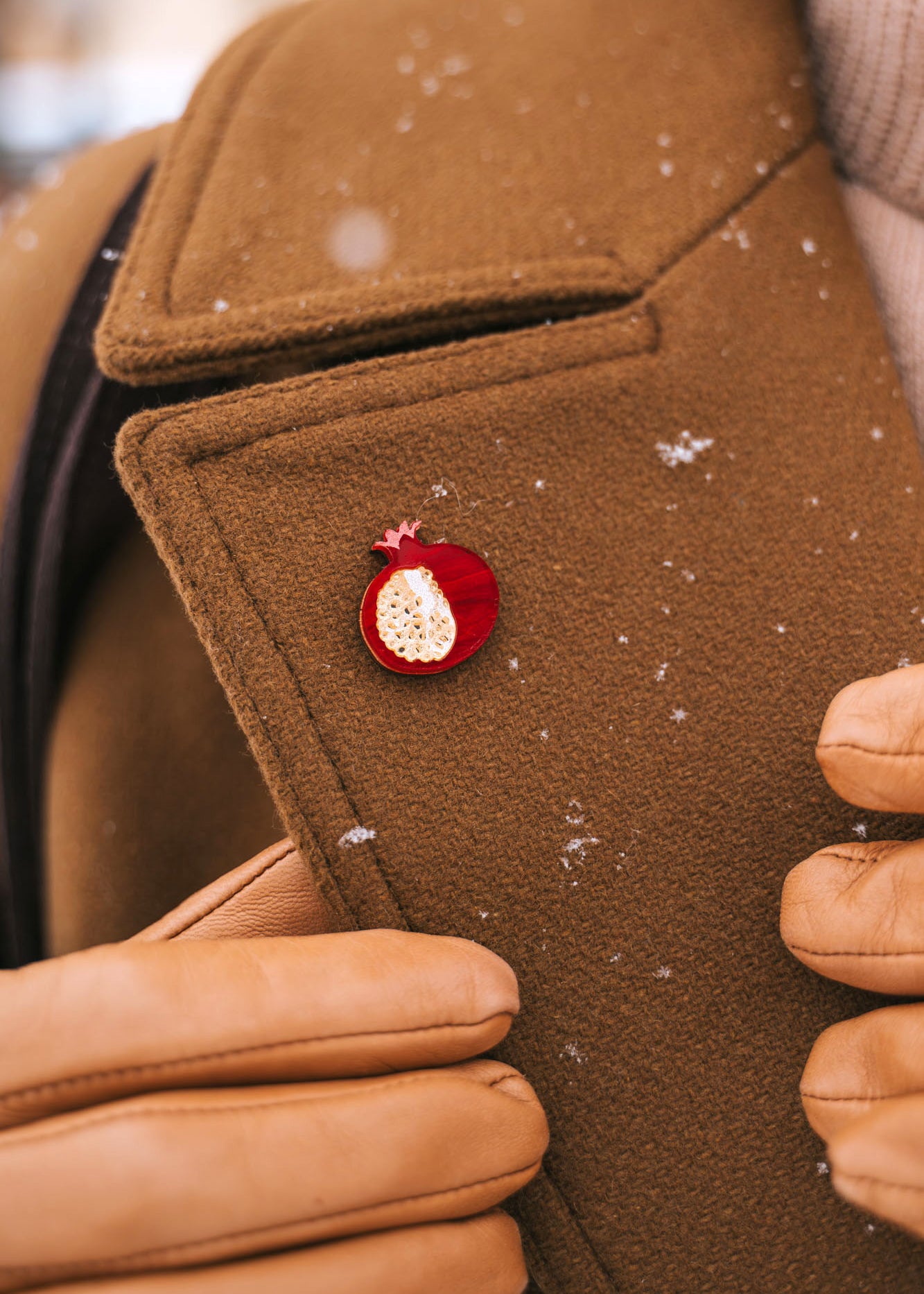 A person wearing a brown coat and tan gloves adjusts the lapel, which is decorated with a handcrafted Pomegranate Pin—a unique fashion accessory. Snowflakes are visible on the coat.