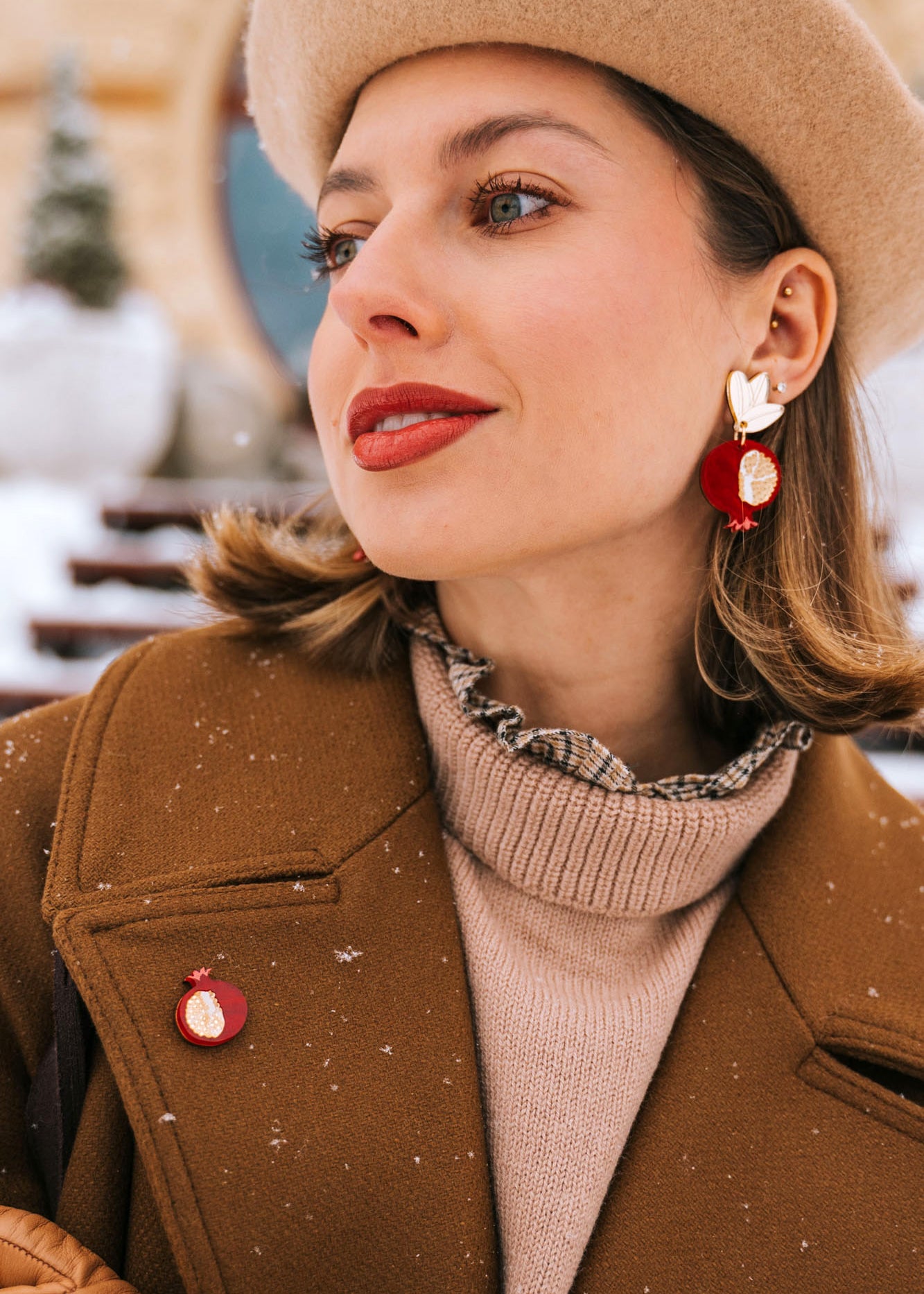 A stylish woman in a beige beret, brown coat, and tan gloves poses outdoors in snowy weather. She wears red and gold handcrafted earrings and a matching Pomegranate Pin fashion accessory, with snowflakes visible on her coat.