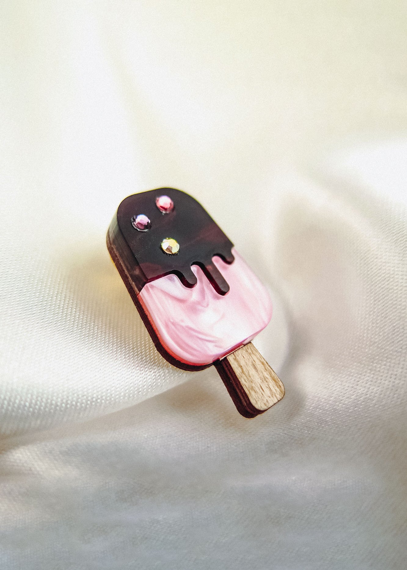 A handcrafted popsicle pin shaped like a pink ice cream bar with a chocolate coating, topped with three small rhinestones, lies on a soft, cream-colored fabric background.