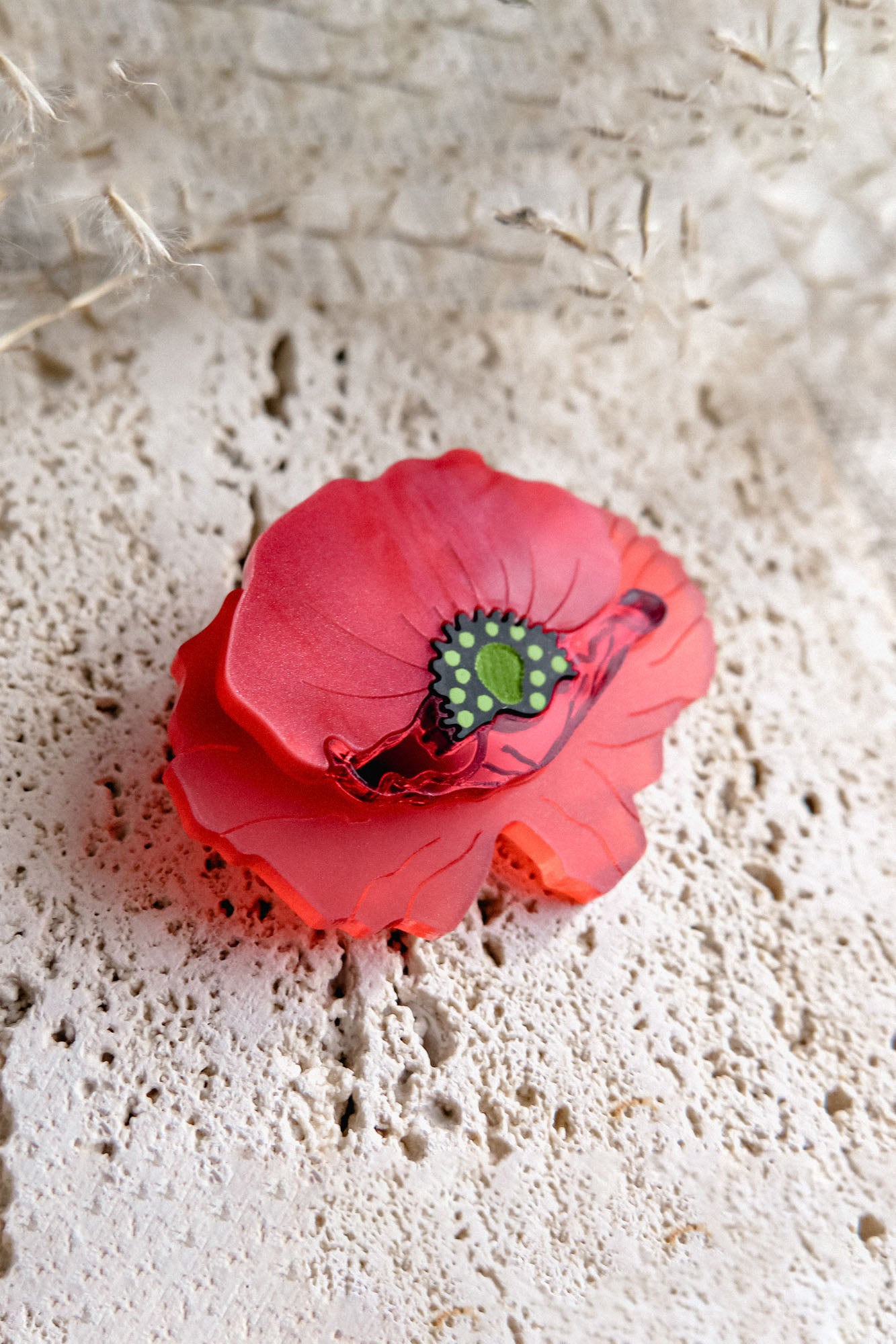 A detailed, red brooch shaped like a poppy with a green and black center rests on a textured, light-colored stone surface, its acrylic glass petals catching the light amid delicate dried grass in the background.
