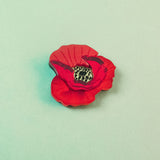 A handmade Red Poppy Flower Lapel Pin with a black and yellow center lies on a pale green background—perfect for fashion enthusiasts seeking a unique accessory.