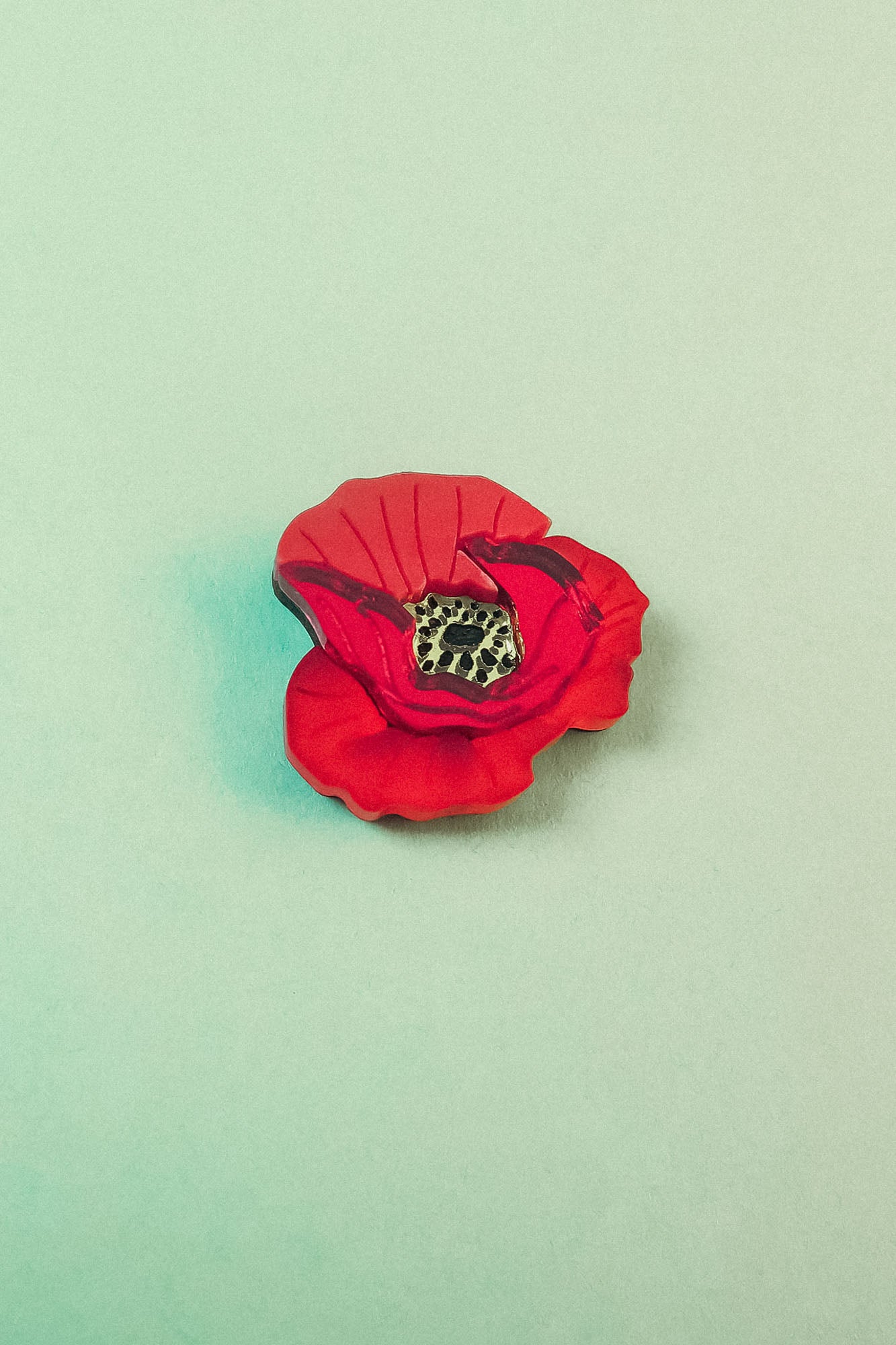 A handmade Red Poppy Flower Lapel Pin with a black and yellow center lies on a pale green background—perfect for fashion enthusiasts seeking a unique accessory.