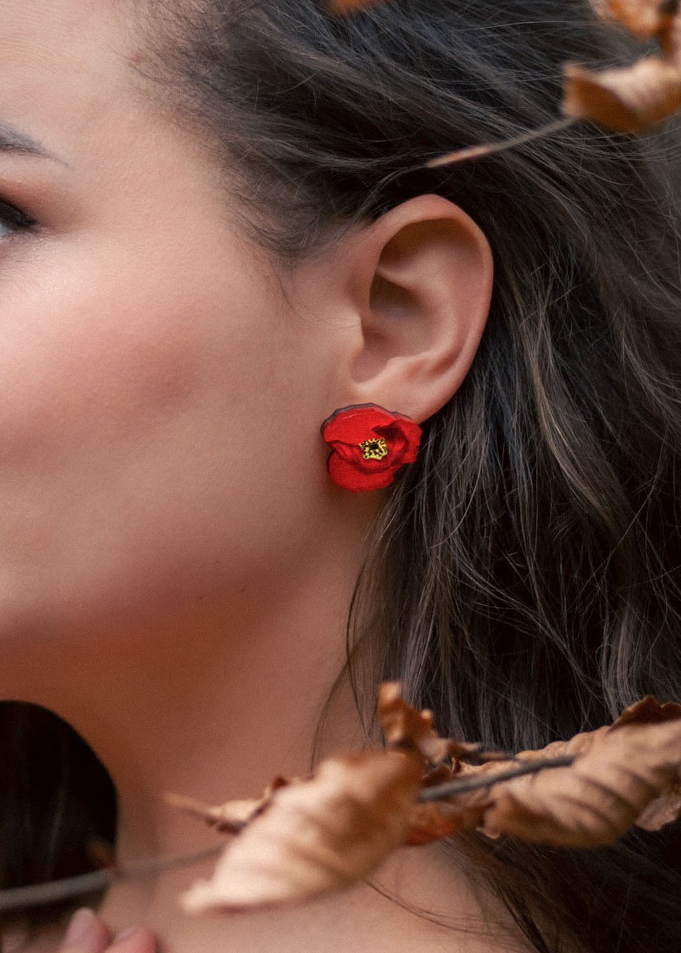 Close-up of a womans ear adorned with a bright red poppy design acrylic earring. Part of her face and dark, wavy hair are visible, with dried brown leaves in the foreground. The earring is lightweight and eye-catching.
