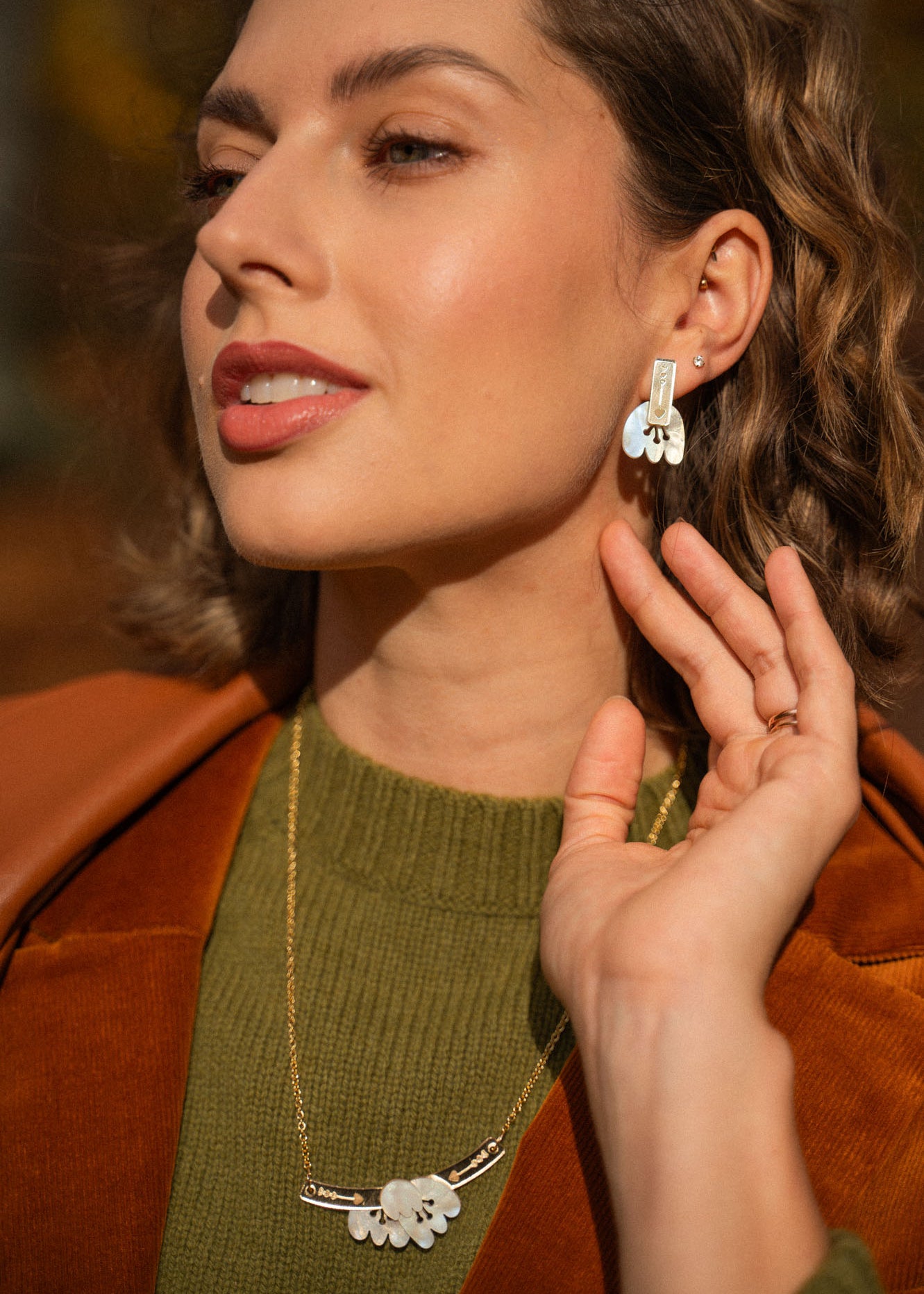 A woman with wavy, shoulder-length hair wears a green sweater, brown jacket, and gold handcrafted earrings—Romantic Tulip Earrings—with a matching necklace, gently touching her ear and smiling softly in warm sunlight.