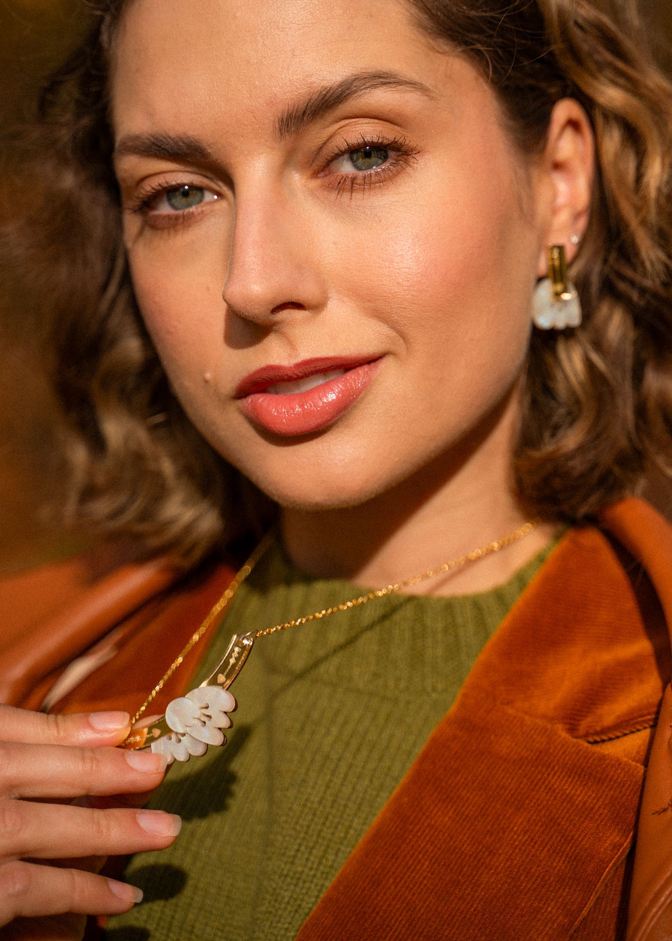 A woman with wavy brown hair wears an elegant green top, brown jacket, and handcrafted gold jewelry with matching white flower-shaped earrings and a Tulip Necklace, holding the necklace and looking confidently at the camera.