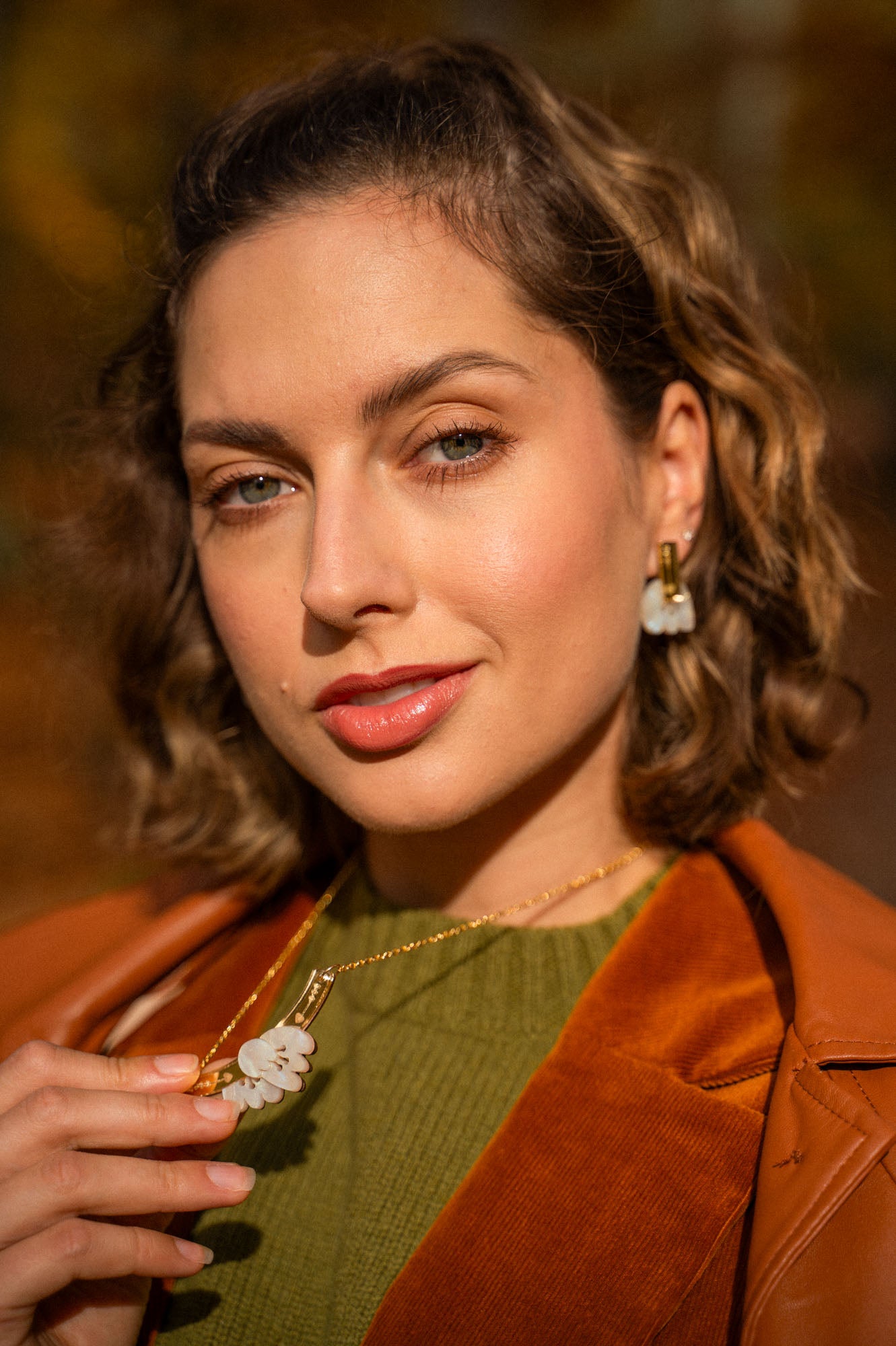 A woman with wavy brown hair wears an elegant green top, brown jacket, and handcrafted gold jewelry with matching white flower-shaped earrings and a Tulip Necklace, holding the necklace and looking confidently at the camera.