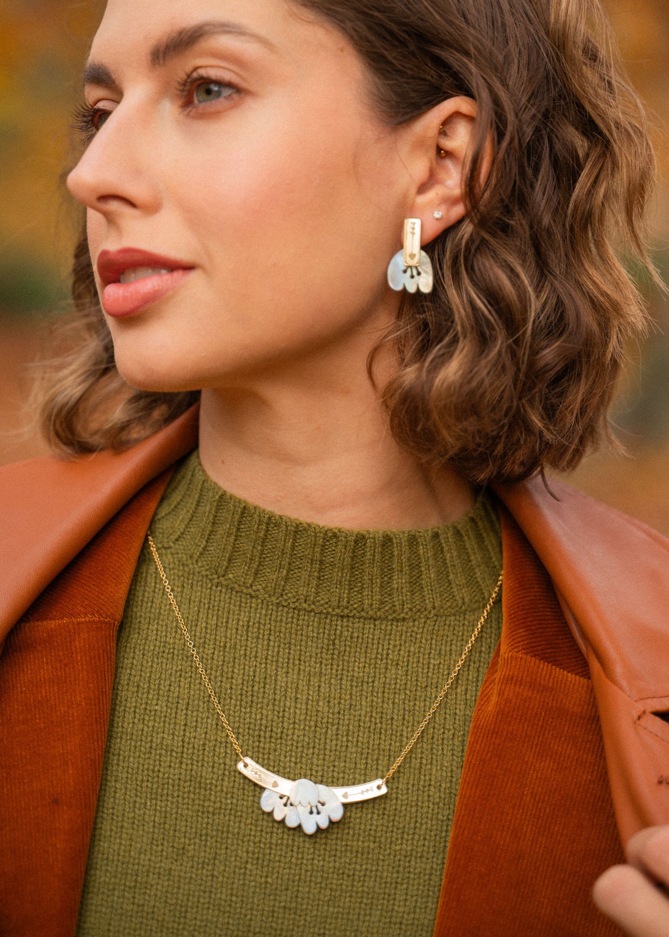 A woman with short wavy brown hair wears a green sweater, brown jacket, and an elegant Tulip Necklace with matching earrings featuring white accents, standing outdoors with blurred autumn foliage in the background.