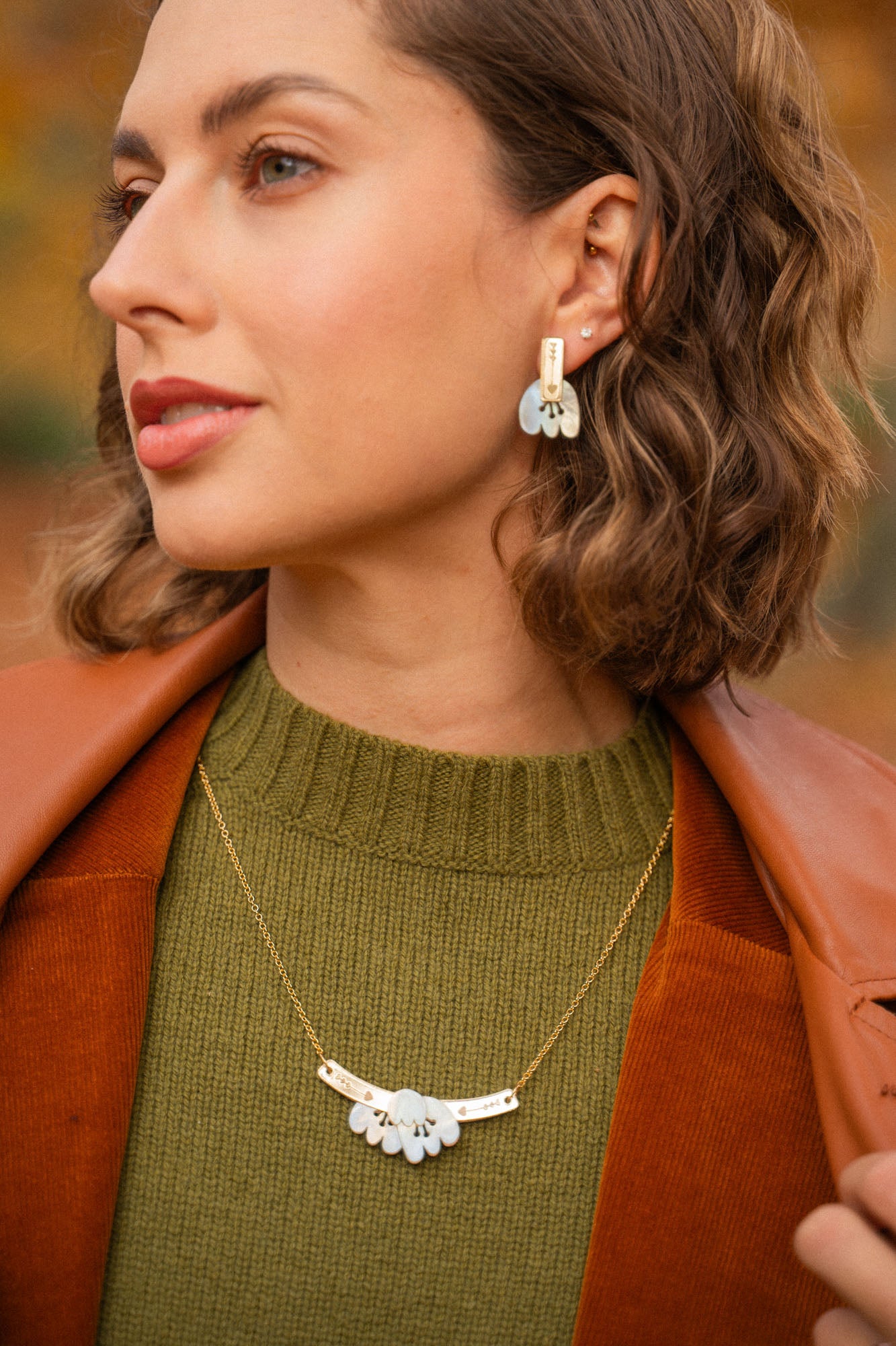 A woman with short wavy brown hair wears a green sweater, brown jacket, and an elegant Tulip Necklace with matching earrings featuring white accents, standing outdoors with blurred autumn foliage in the background.