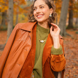 A woman smiles elegantly while standing in a forest with autumn leaves. She wears a brown leather coat over an orange suede jacket, with a green sweater, patterned skirt, and a handcrafted Tulip Necklace. Tall trees and fallen leaves fill the background.