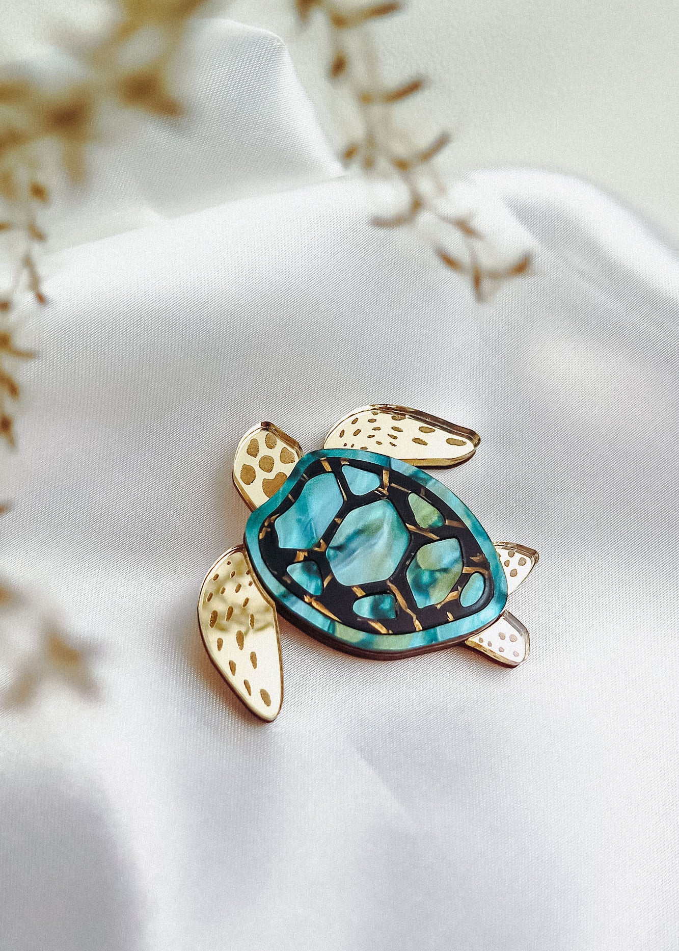 A handcrafted sea turtle brooch with a gold and turquoise mosaic shell rests on smooth white fabric. Its fins are gold with brown spots, while delicate dried flowers blur the foreground, highlighting its artistic design.