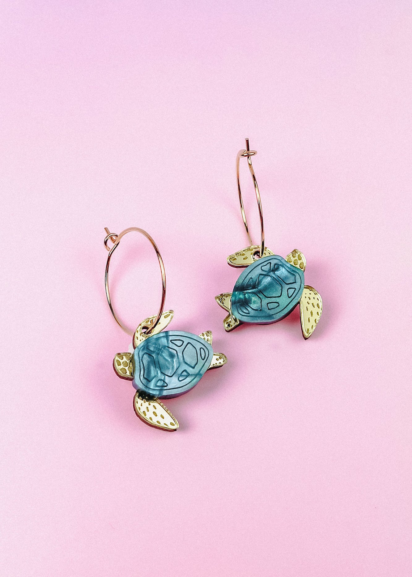 Handcrafted Little Turtle Hoop Earrings featuring gold hoops and blue and gold sea turtle charms, displayed on a pink background.