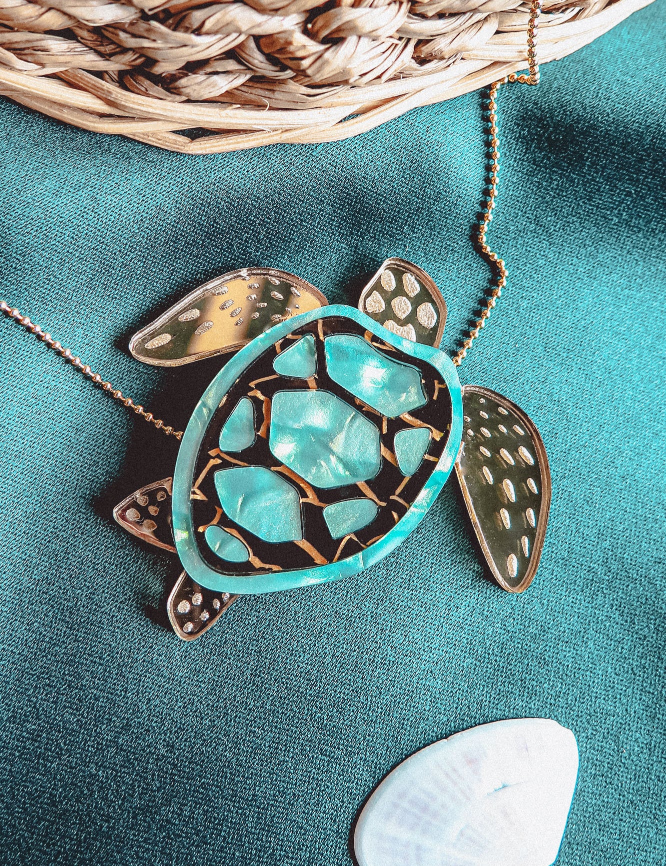 A close-up of a handcrafted Sea Turtle Necklace pendant with a turquoise and gold shell, lying on green fabric next to a white seashell and a wicker basket.