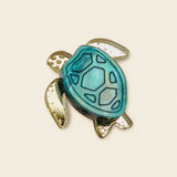 A handcrafted sea turtle pin shaped like a turtle, featuring a blue and teal shell with gold-colored flippers and outline, made from mirror-acrylic glass and set against a plain beige background.