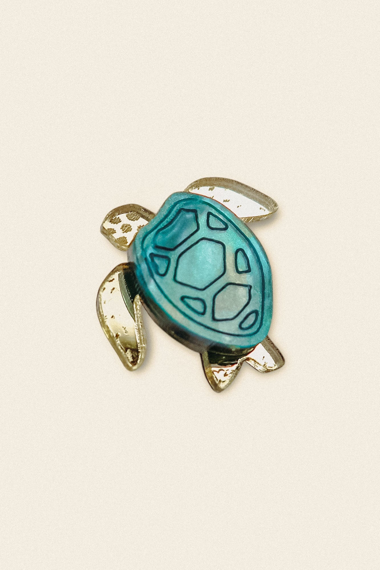 A handcrafted sea turtle pin shaped like a turtle, featuring a blue and teal shell with gold-colored flippers and outline, made from mirror-acrylic glass and set against a plain beige background.