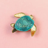 A small handcrafted sea turtle pin with a blue-green shell and gold-colored flippers, pictured on a pink background.